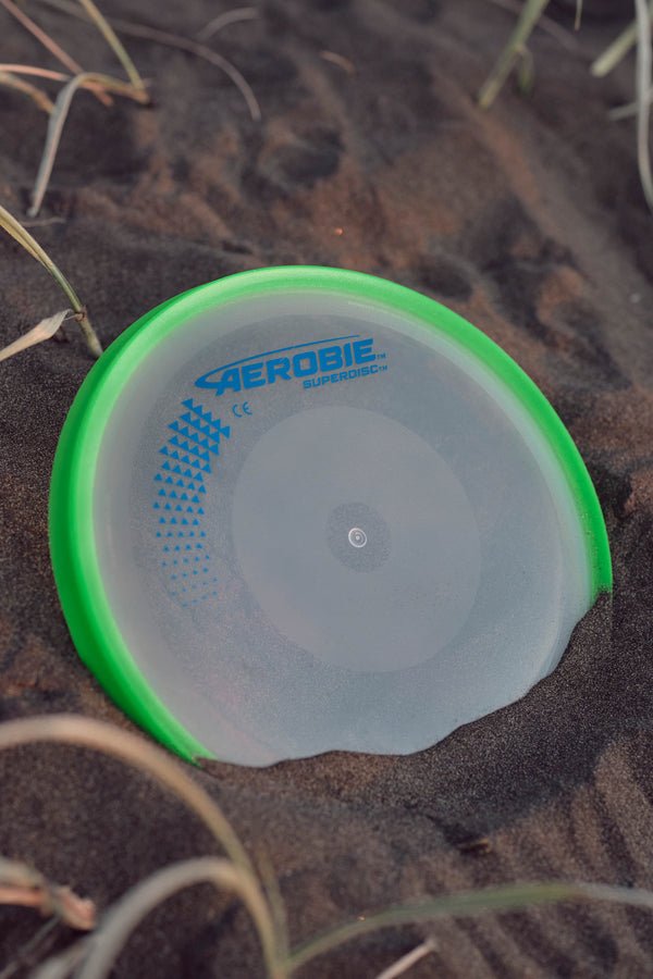 The Aerobie Superdisc by Other, a flying disc with a green rim, is partly buried in dark sand with a few blades of grass around it.