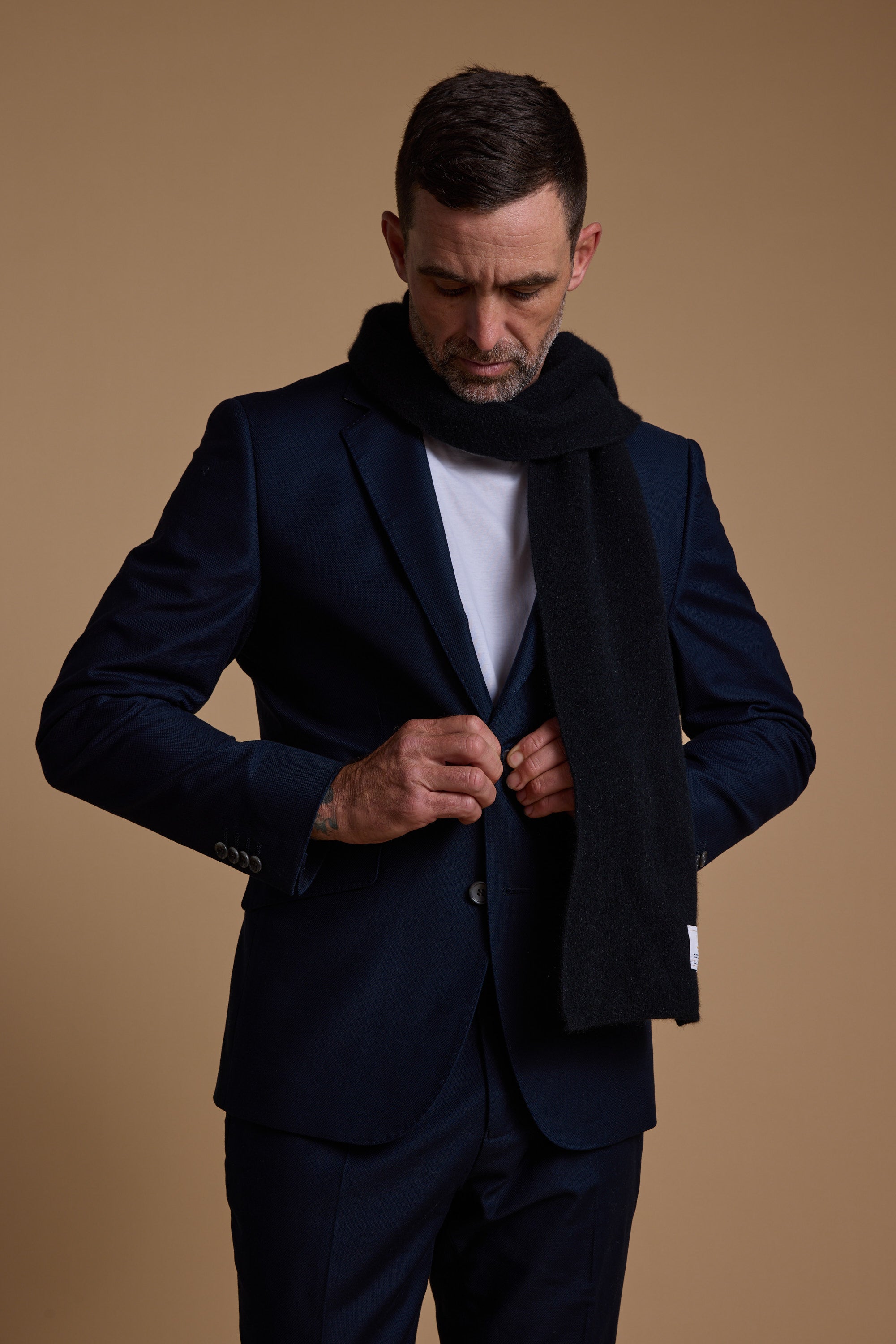 A man in a navy suit and white shirt wears a Woolyarns Perino NZ Made Scarf, crafted from a luxurious merino, possum, and silk blend, as he buttons his jacket against a plain tan background.