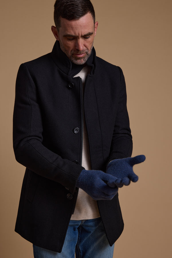A man in a black coat and white shirt adjusts his NZ Made Perino Gloves by Woolyarns Perino, crafted from a merino possum silk blend. He stands against a plain beige background, also wearing blue jeans.