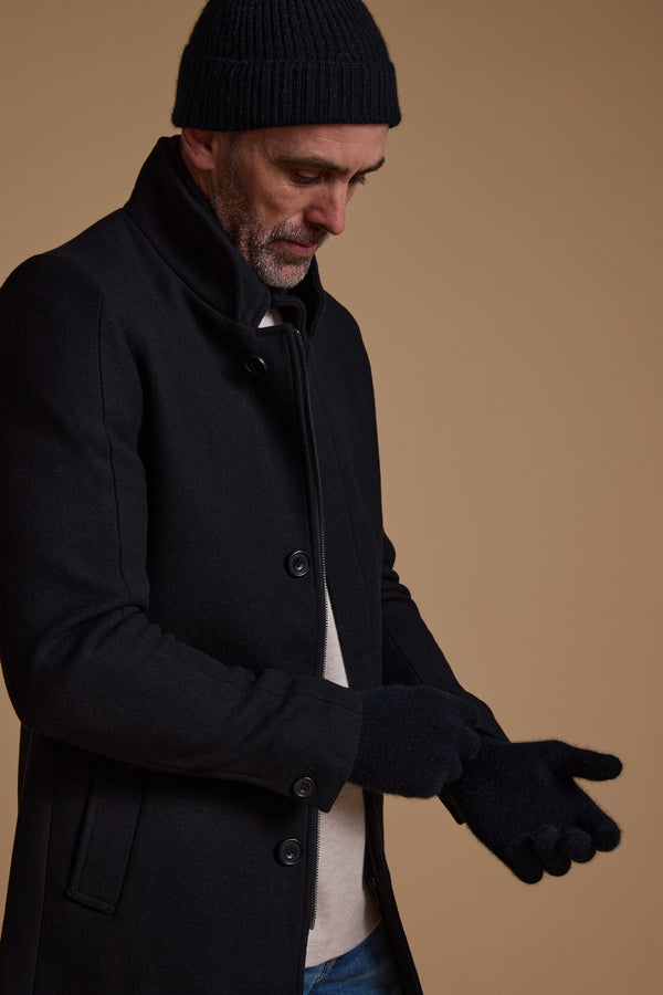 A man in a black coat and knit hat adjusts his NZ Made Perino Gloves by Woolyarns Perino, standing against a plain brown background.