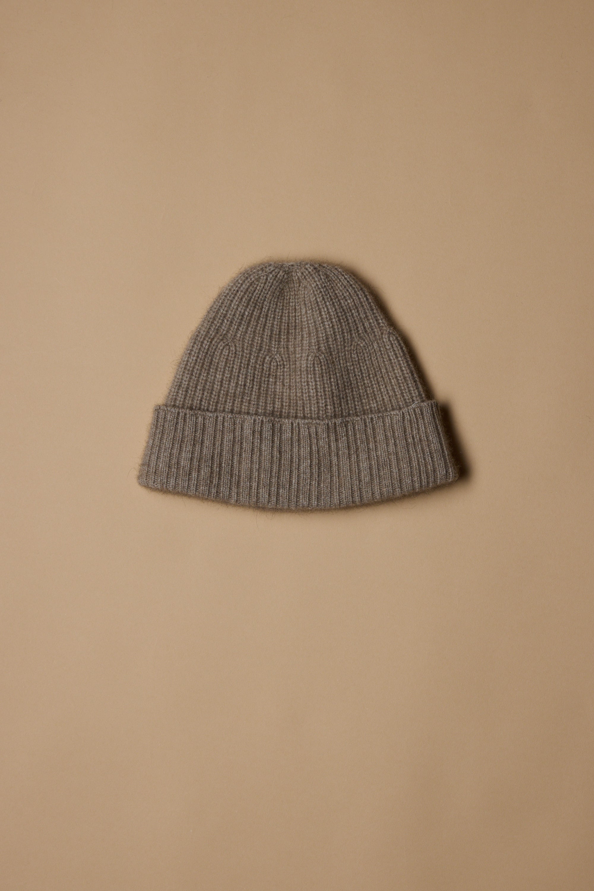 The Woolyarns Perino NZ Made Perino Beanie, in gray and crafted from a luxurious merino possum silk blend, features a folded brim and is displayed on a plain light brown background.