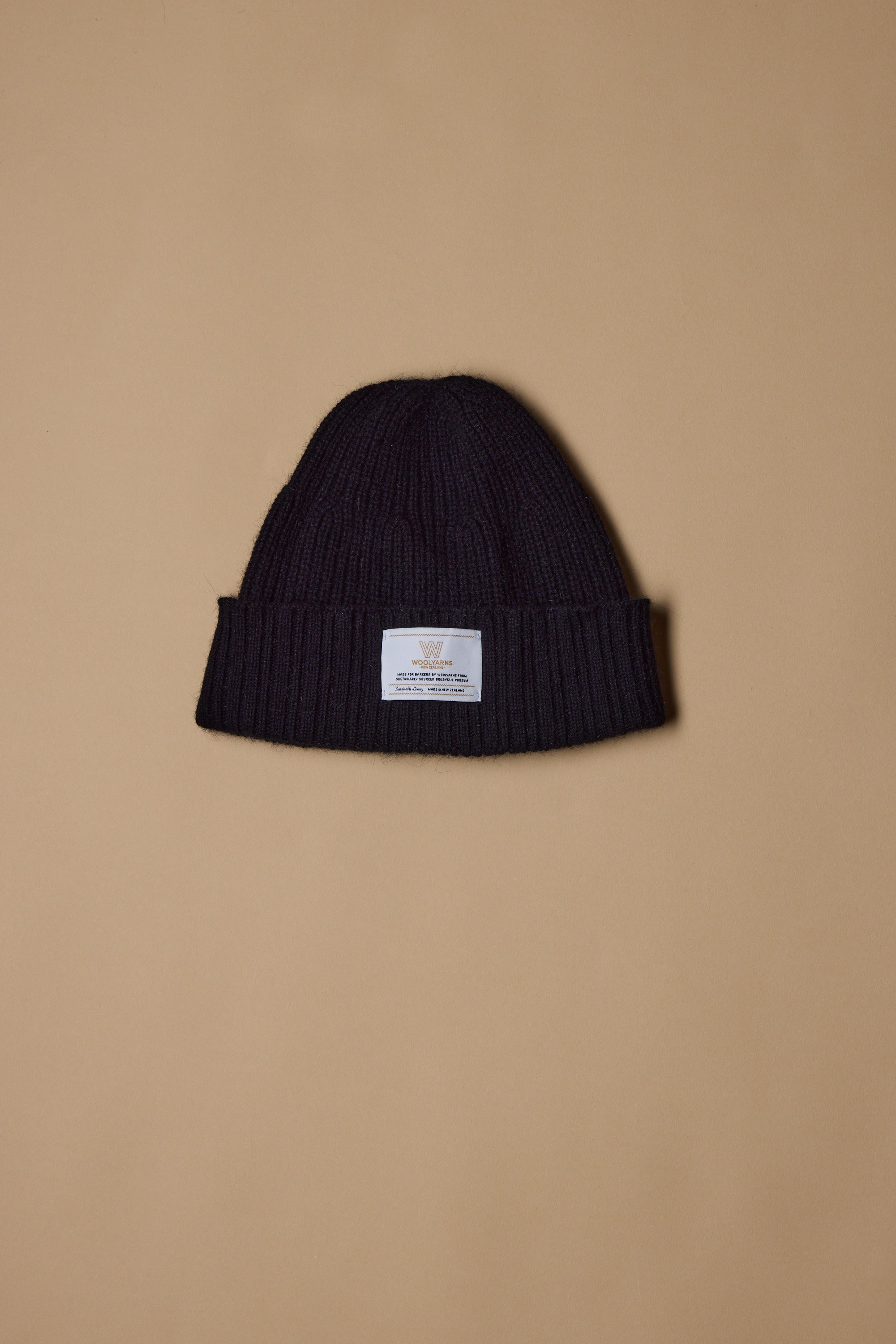 The NZ Made Perino Beanie by Woolyarns Perino is a black knit merino possum silk beanie with a folded brim and white front label, shown on a beige background. It belongs to the premium Perino accessories collection.