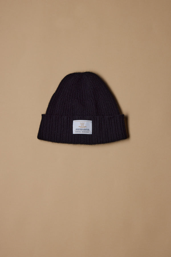 The NZ Made Perino Beanie by Woolyarns Perino is a black knit merino possum silk beanie with a folded brim and white front label, shown on a beige background. It belongs to the premium Perino accessories collection.