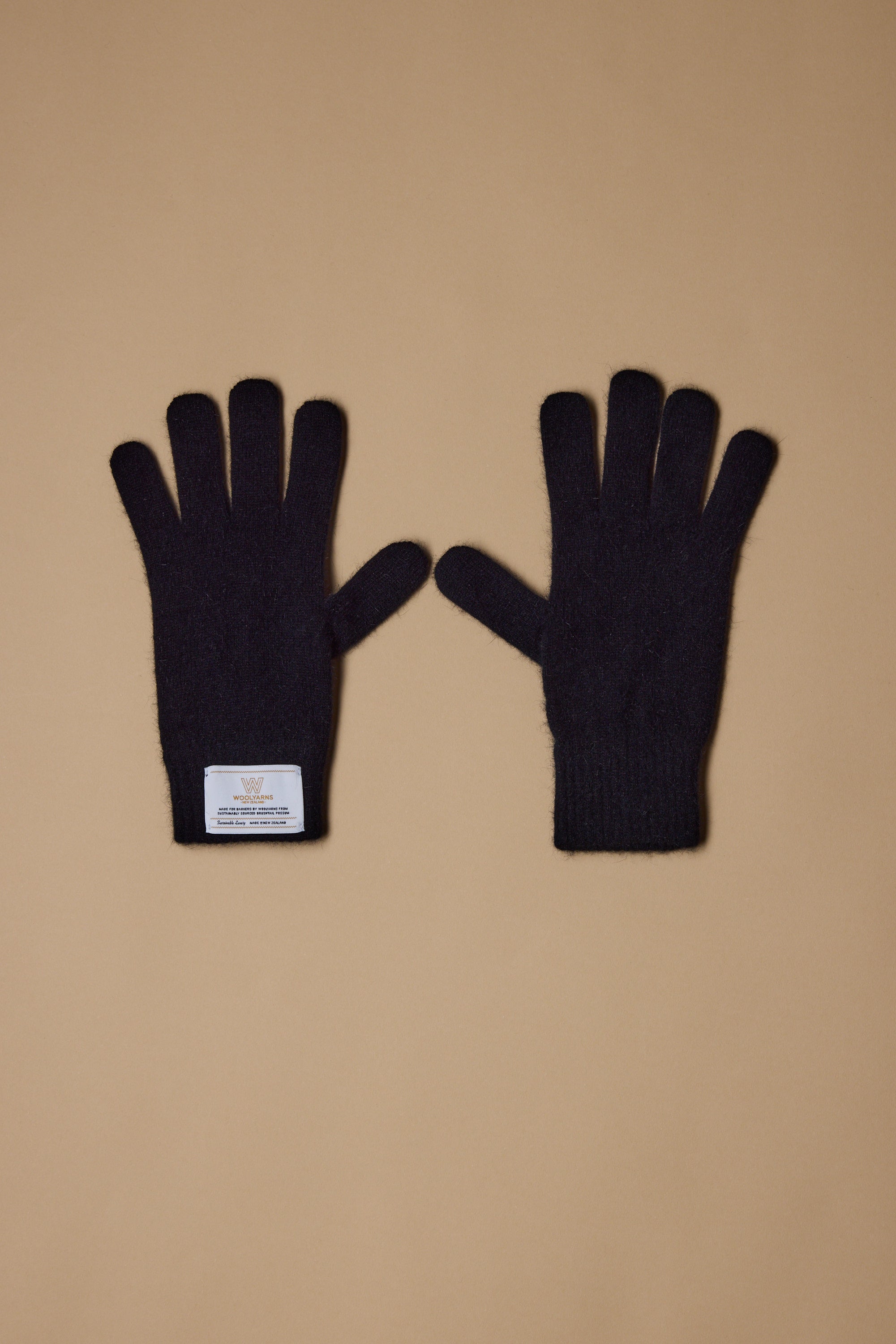 A pair of black NZ Made Perino Gloves by Woolyarns Perino, crafted from a merino possum silk blend, lay flat on a light brown surface with a white label sewn onto the wrist of the left glove. Made in New Zealand.