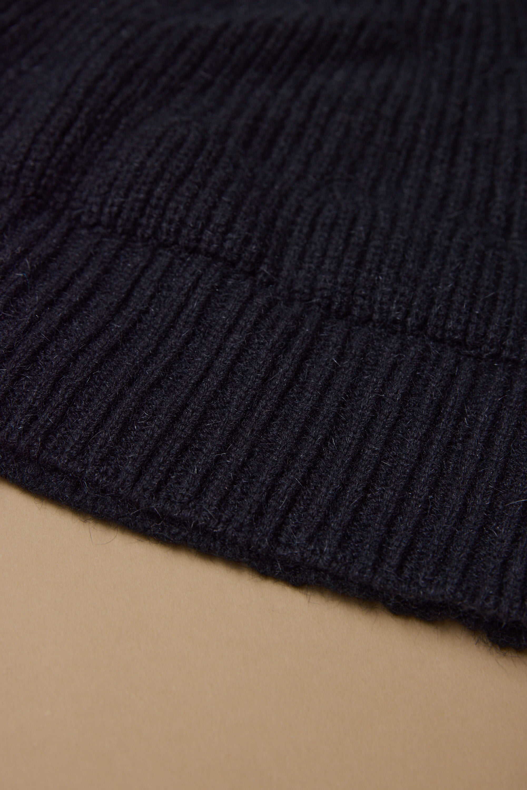 Close-up of a black ribbed knit fabric from the Woolyarns Perino NZ Made Perino Beanie, laid on a light brown surface. The texture and stitching detail highlight the premium quality of New Zealand made merino possum silk.