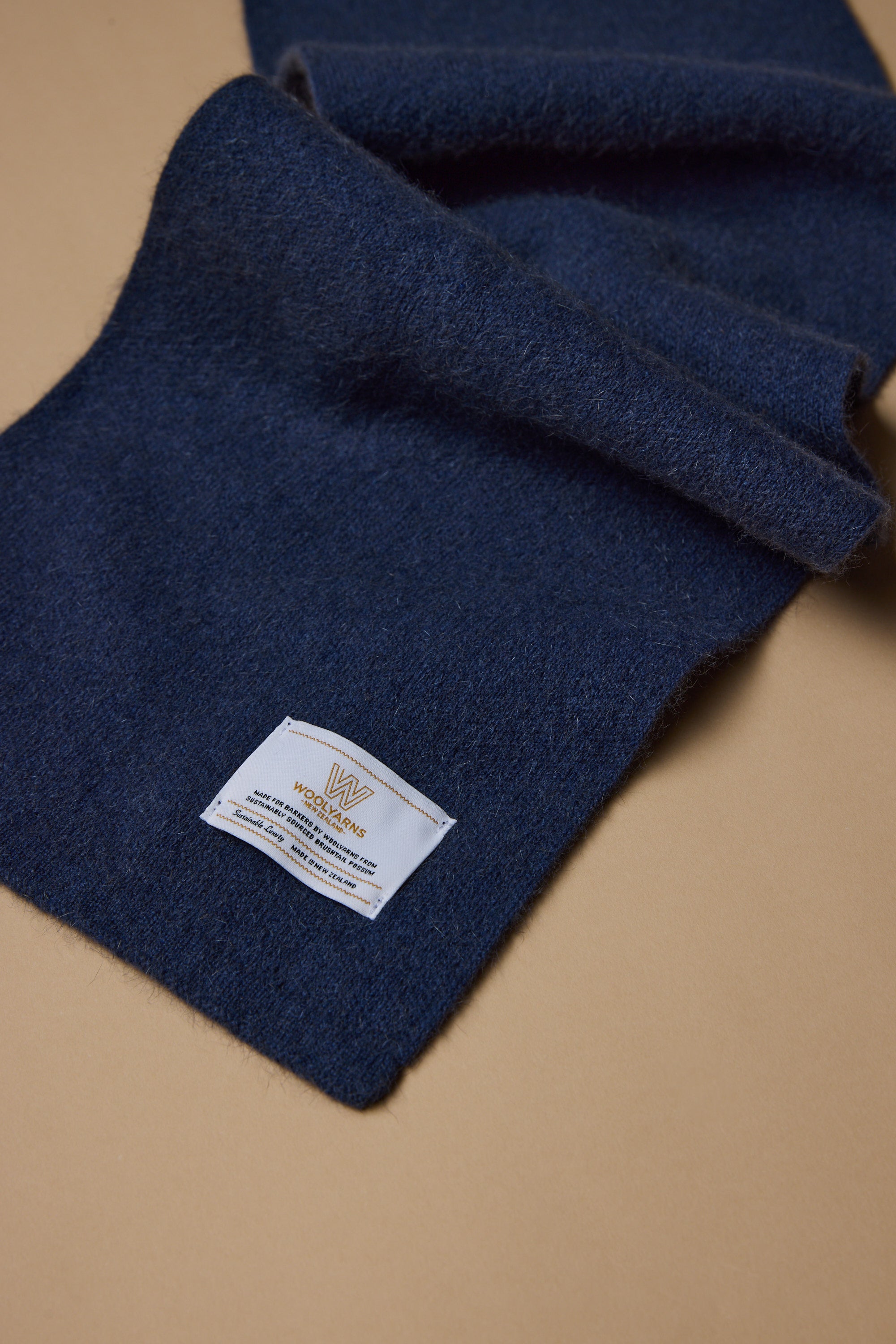 The NZ Made Perino Scarf by Woolyarns Perino is a dark blue merino possum silk blend, with a white fabric label in gold and blue text, displayed on a light brown surface. Crafted sustainably in New Zealand as part of the Perino accessories range.