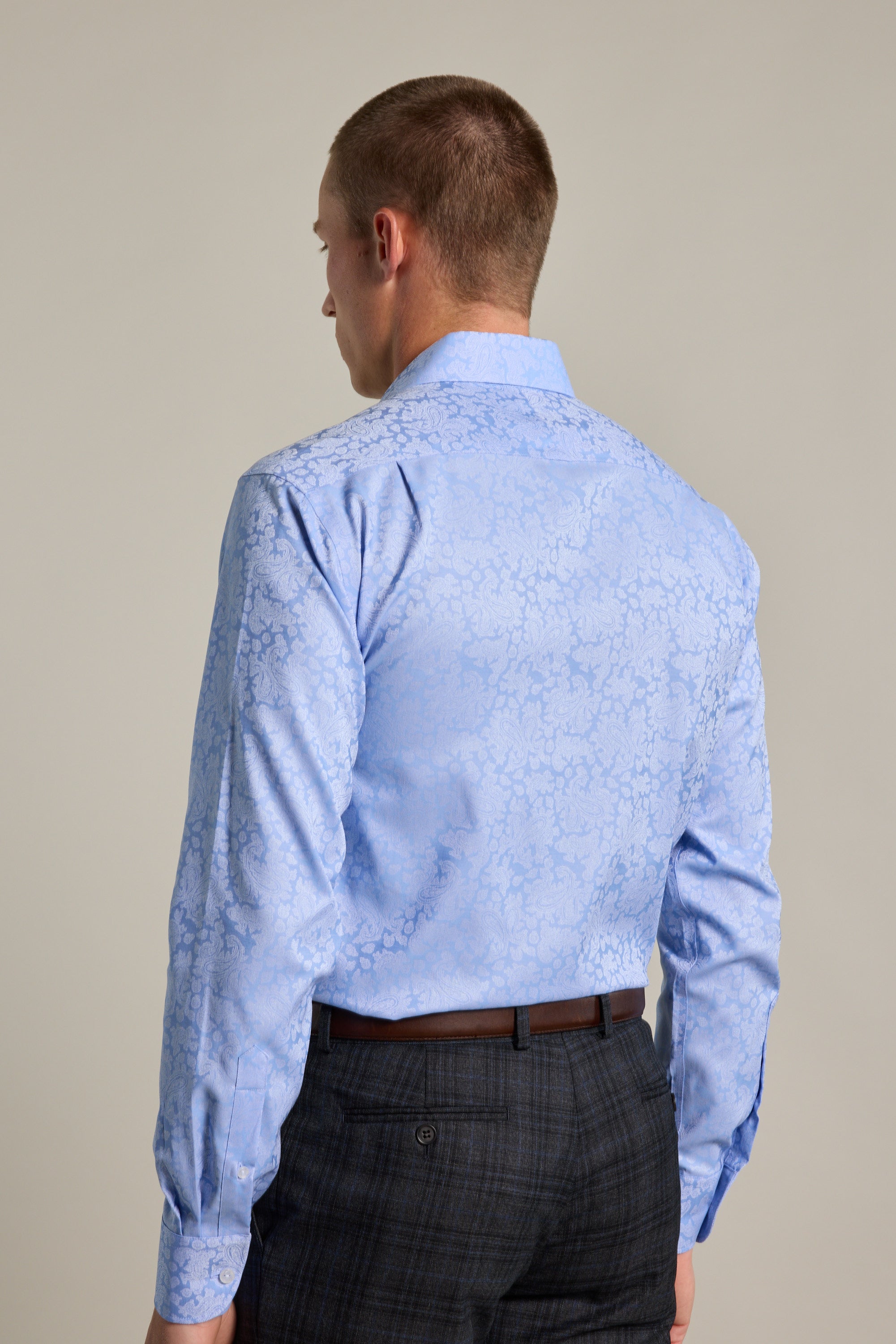 A man is seen from behind wearing the Barkers Corran Paisley Business Shirt in light blue with a subtle pattern, paired with gray plaid trousers. The background is plain and neutral.