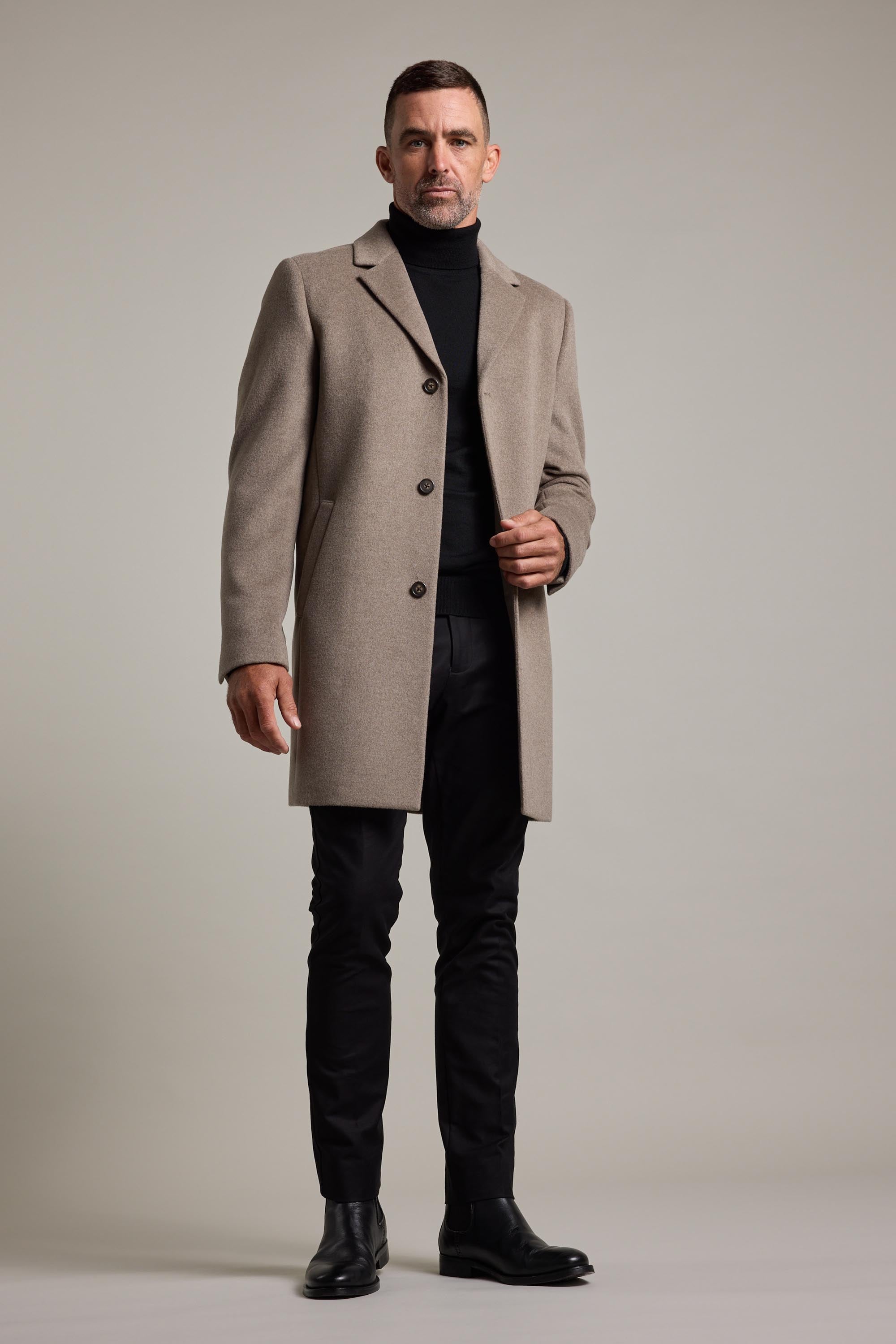 A man stands against a plain background wearing the Barkers Morrison Coat, a beige wool blend design, paired with a black turtleneck, pants, and boots. He looks confident and stylish—holding one side of this iconic wardrobe piece.