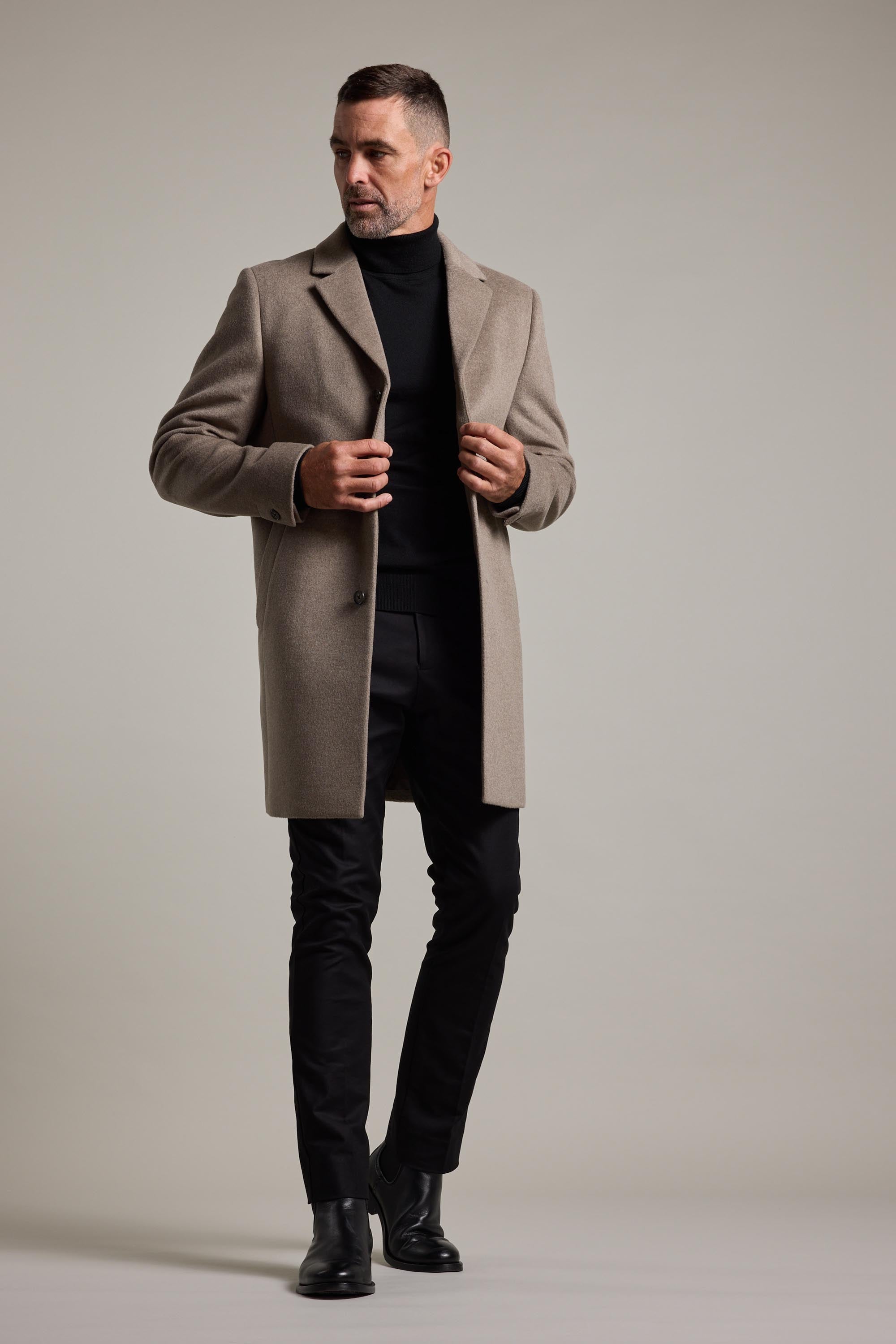 A man poses against a plain background wearing Barkers' light brown wool blend Morrison Coat, paired with a black turtleneck, pants, and boots. He looks to the side, adjusting this wardrobe icon with both hands.
