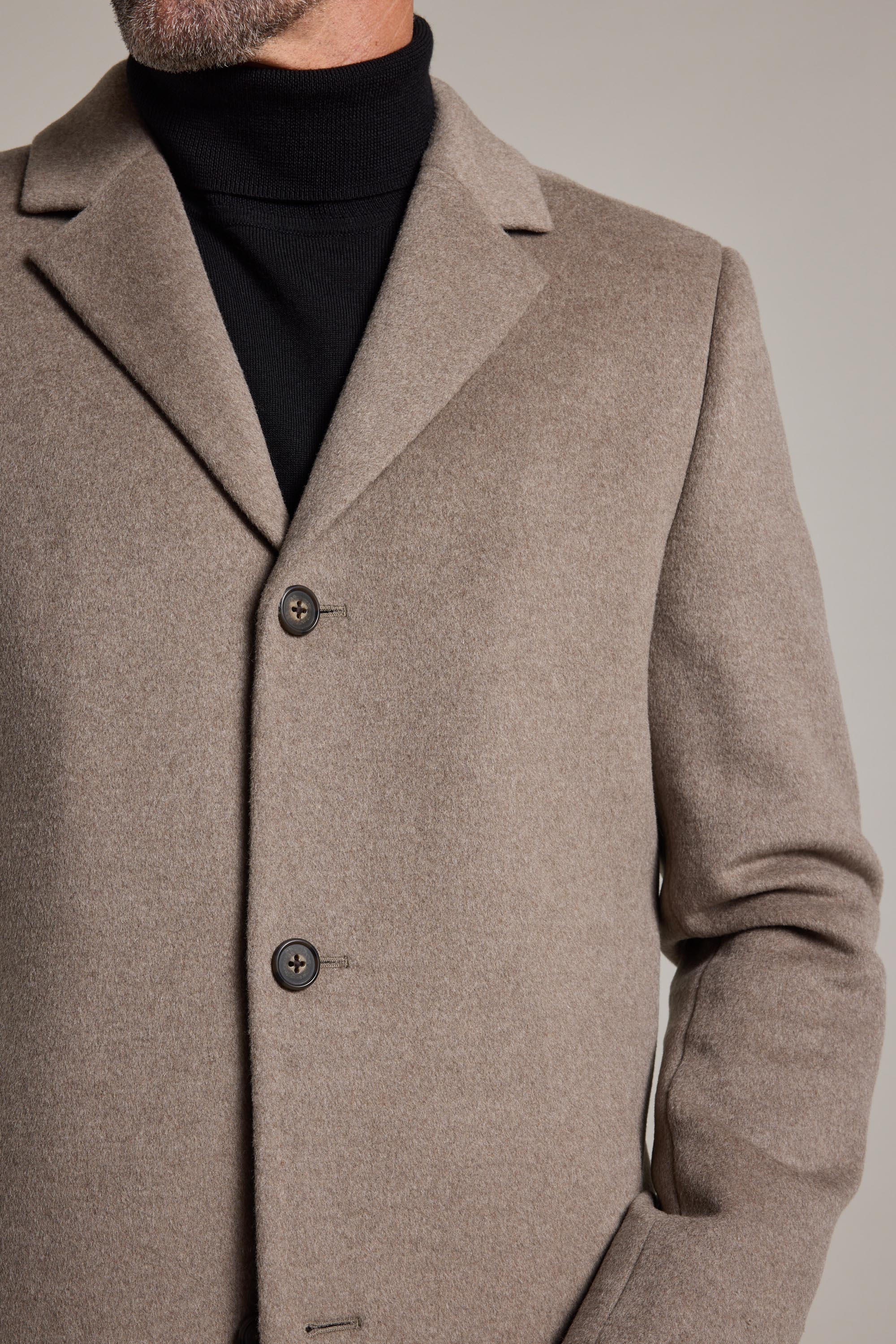 A man, shown from the shoulders down with his left hand in the pocket, wears Barkers’ iconic Morrison Coat—a beige wool-blend coat with dark buttons—over a black turtleneck.