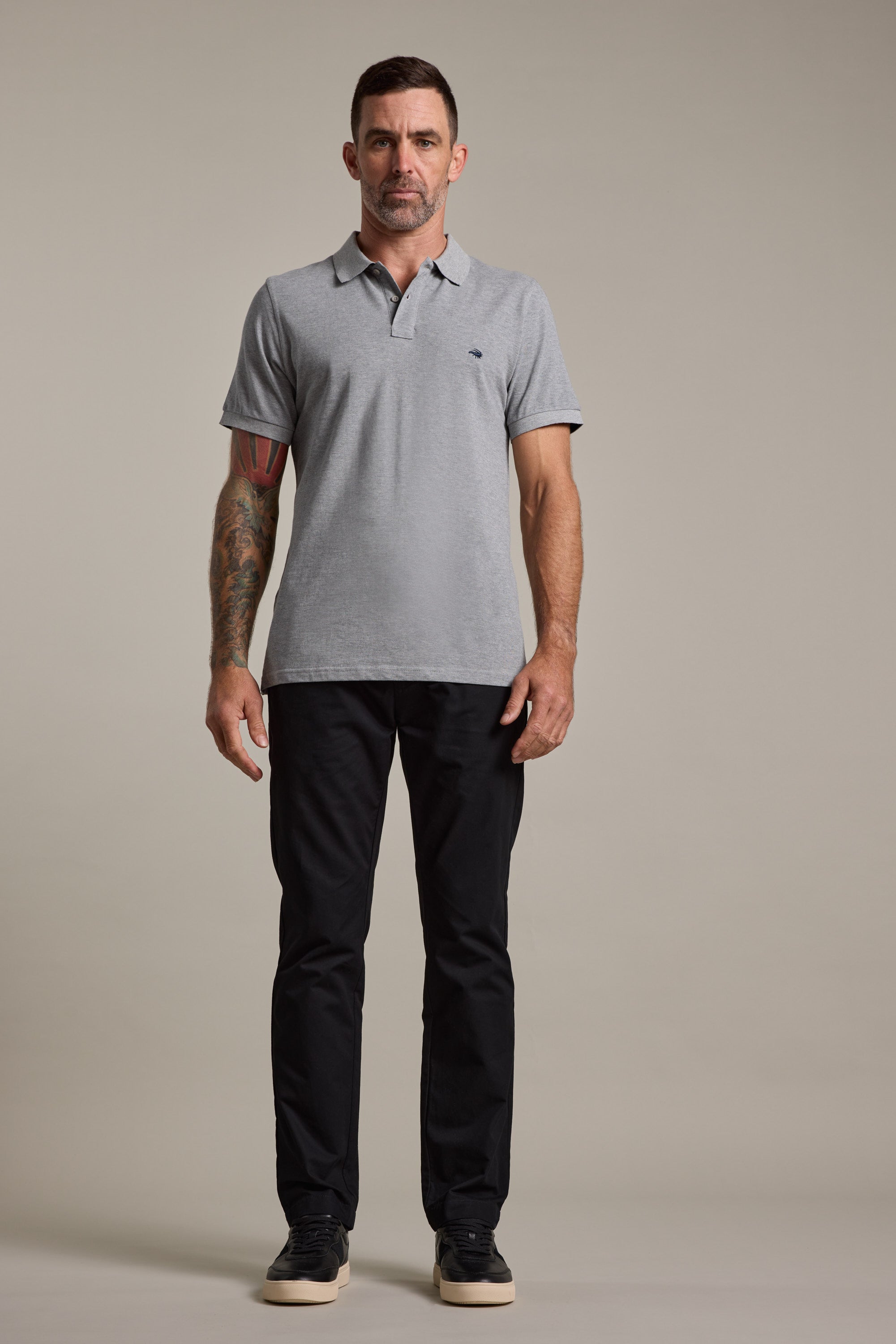 A man with short dark hair and tattoos on his left arm faces forward against a plain background, wearing a light gray polo shirt, black Barkers Dundas Chino trousers, and black sneakers.