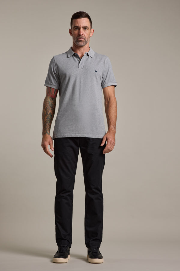 A man with short dark hair and tattoos on his left arm faces forward against a plain background, wearing a light gray polo shirt, black Barkers Dundas Chino trousers, and black sneakers.