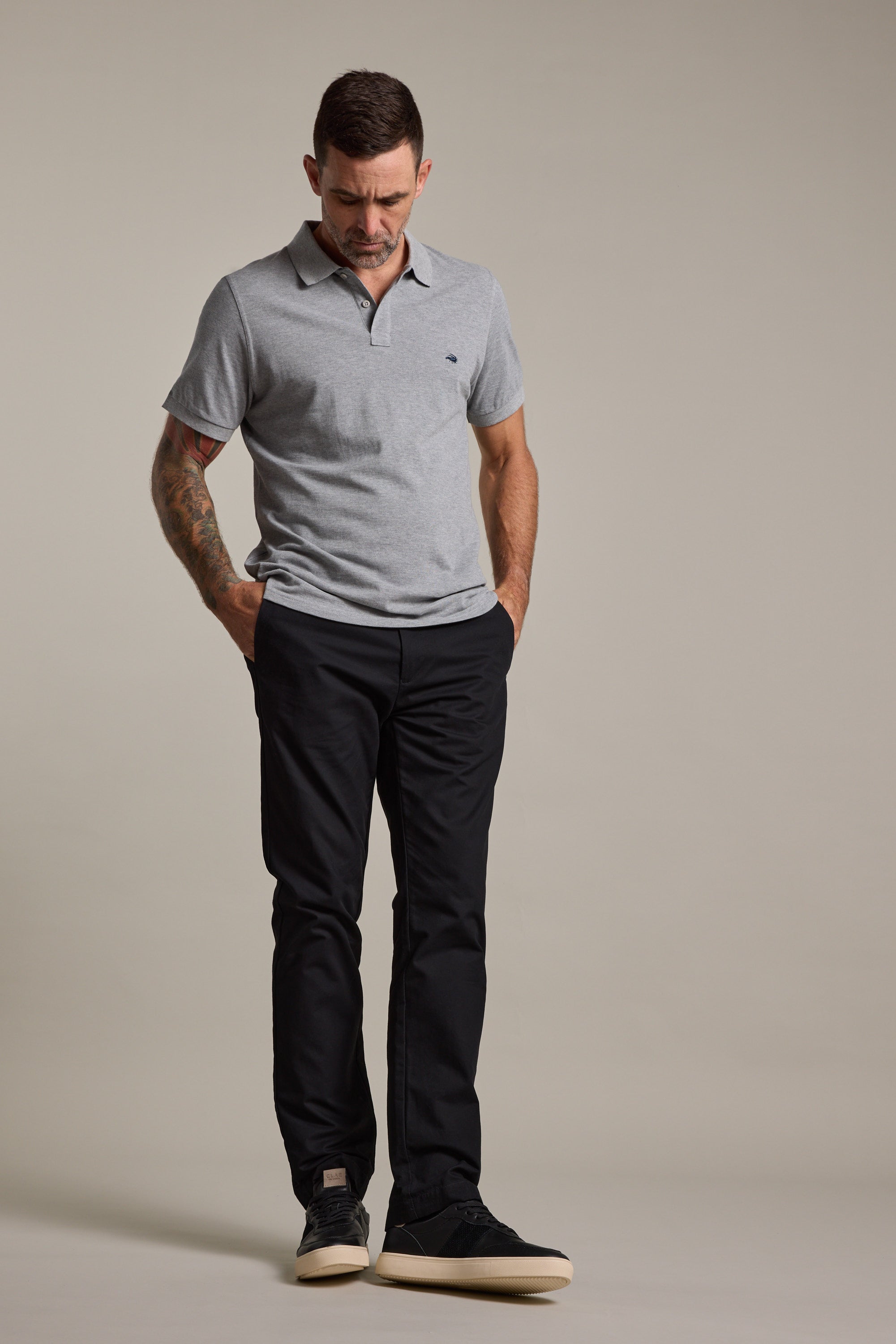 A man with short dark hair, a tattoo on his left arm, wearing a light grey polo shirt and Barkers Dundas Chino, stands with hands in pockets looking down against a plain neutral background. He completes the outfit with black shoes.