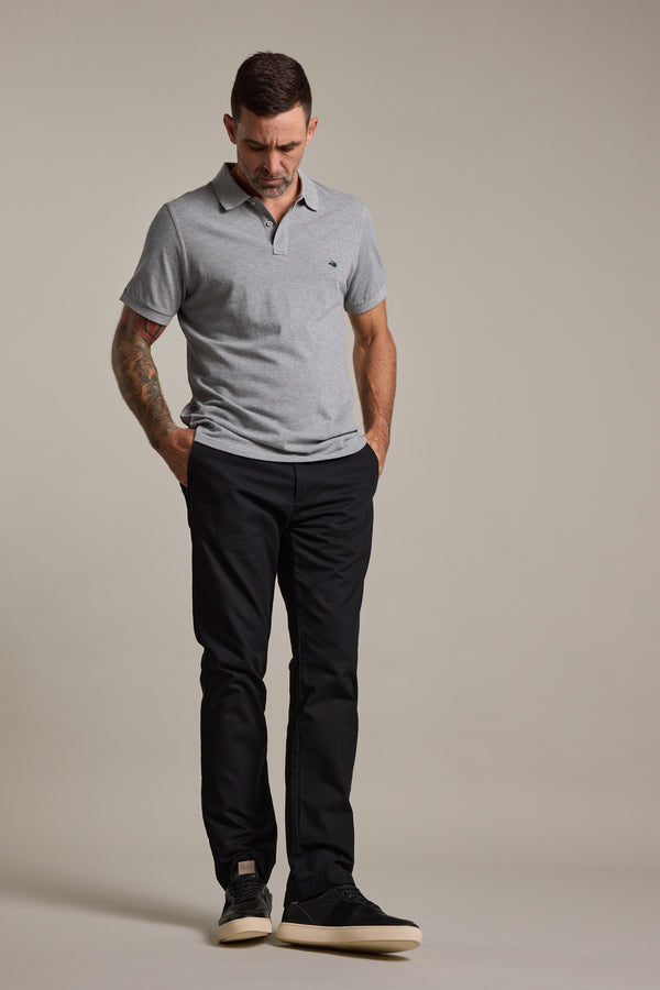 A man with short dark hair, a tattoo on his left arm, wearing a light grey polo shirt and Barkers Dundas Chino, stands with hands in pockets looking down against a plain neutral background. He completes the outfit with black shoes.