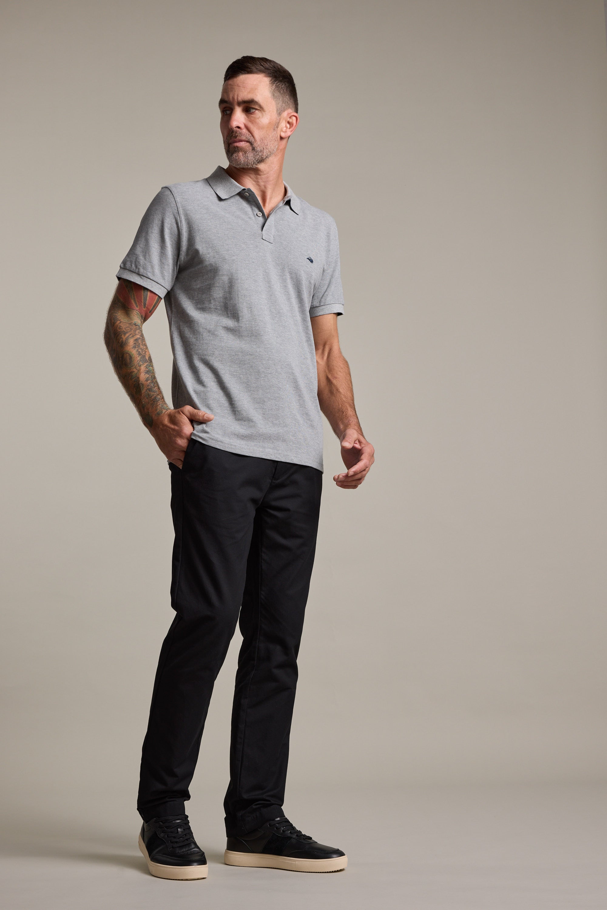 A man with short hair and a tattoo on his right arm stands against a plain background, wearing a light gray polo shirt, Barkers Dundas Chino made from responsibly sourced cotton, and black sneakers with white soles. He looks slightly to the side.