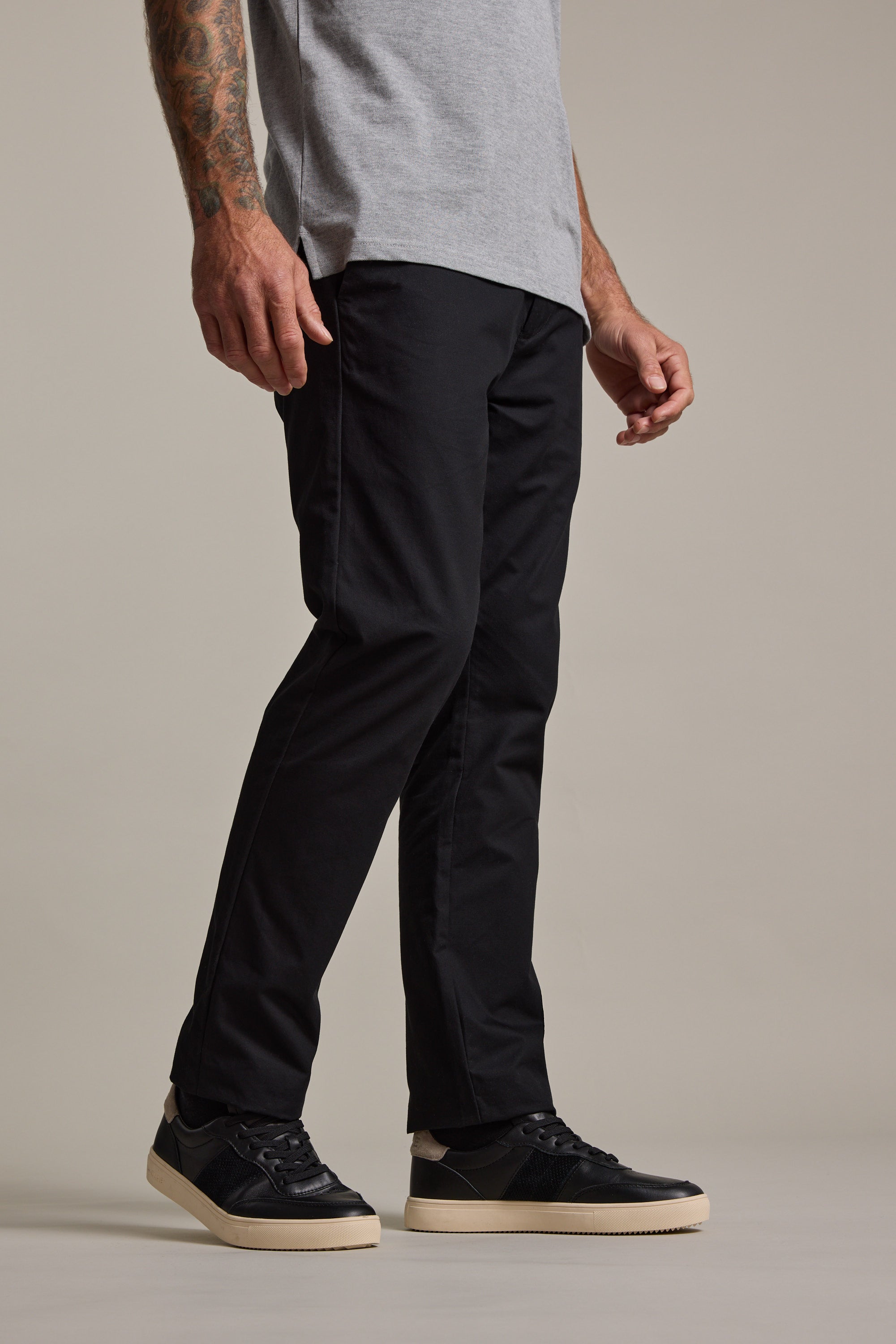 A man, visible from torso to legs, stands against a plain background wearing Barkers Dundas Chino relaxed fit trousers made from responsibly sourced cotton, a light gray T-shirt, and black sneakers with white soles.