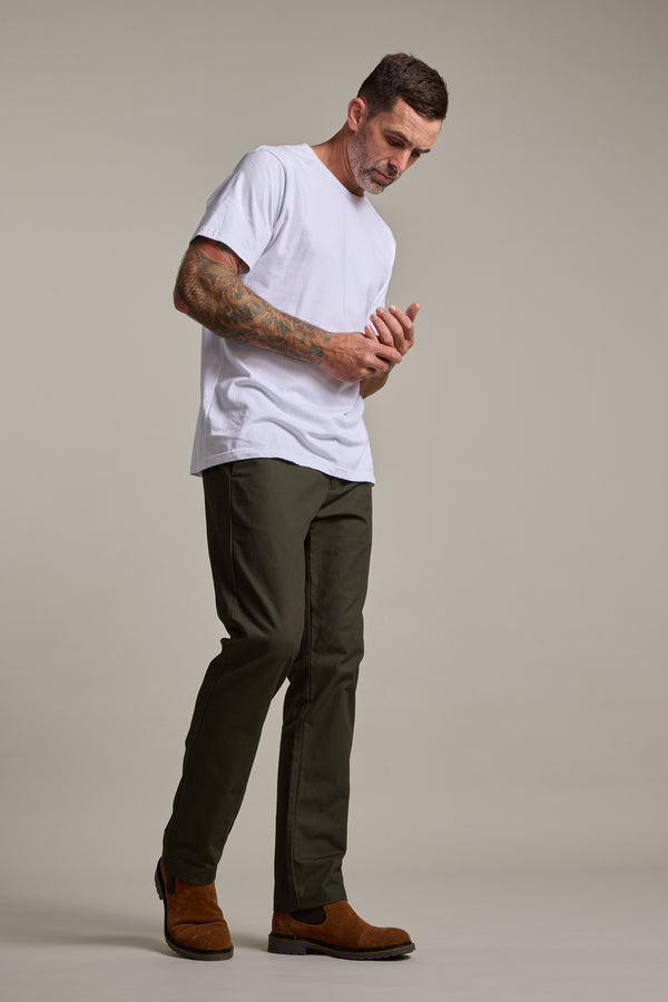 A man with tattoos, wearing a white t-shirt and Barkers Dundas Chino, stands against a plain background, looking down and touching his hands while wearing brown boots.