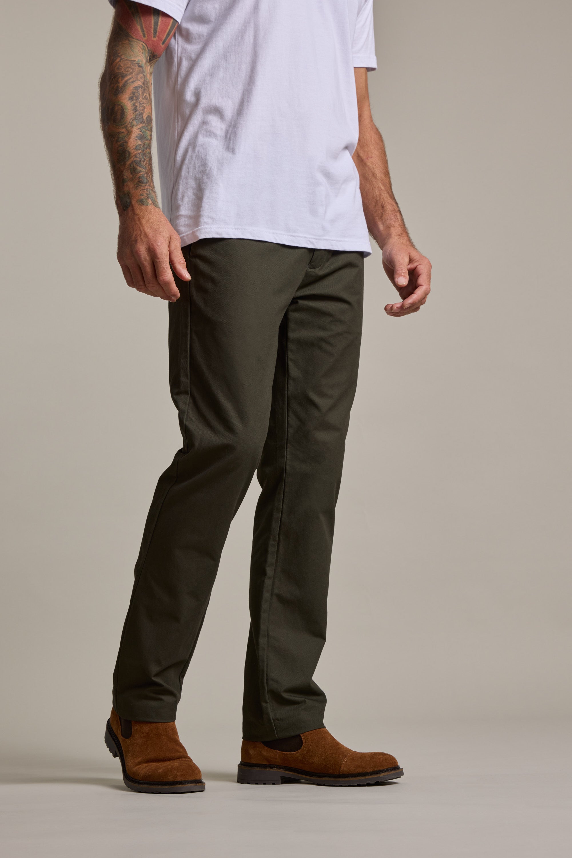 A man stands against a plain background wearing a white t-shirt, Barkers Dundas Chino in relaxed-fit olive green, and brown suede boots. His left arm is tattooed, and his head is out of frame.