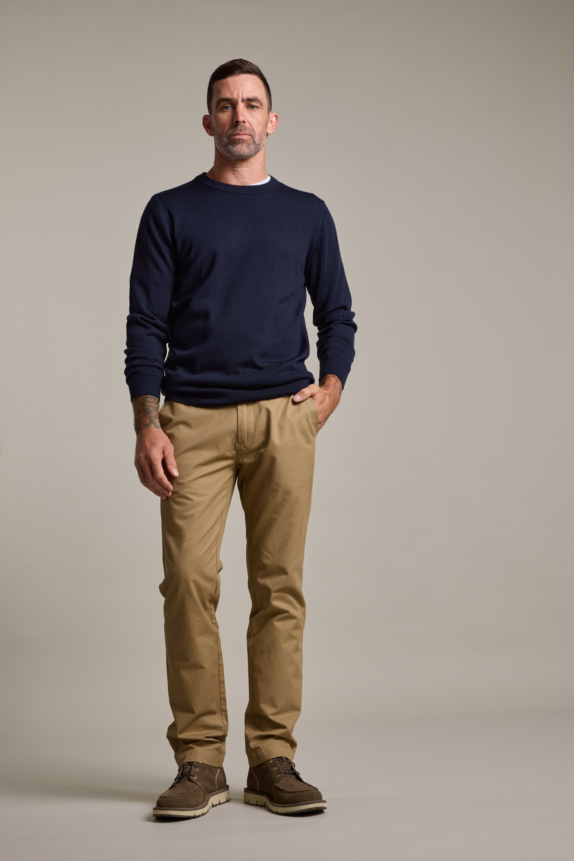 A man with short dark hair and light facial hair stands confidently, wearing Barkers Dundas Chino pants, a navy sweater over a white shirt, and brown shoes against a plain background, with one hand in his pocket.
