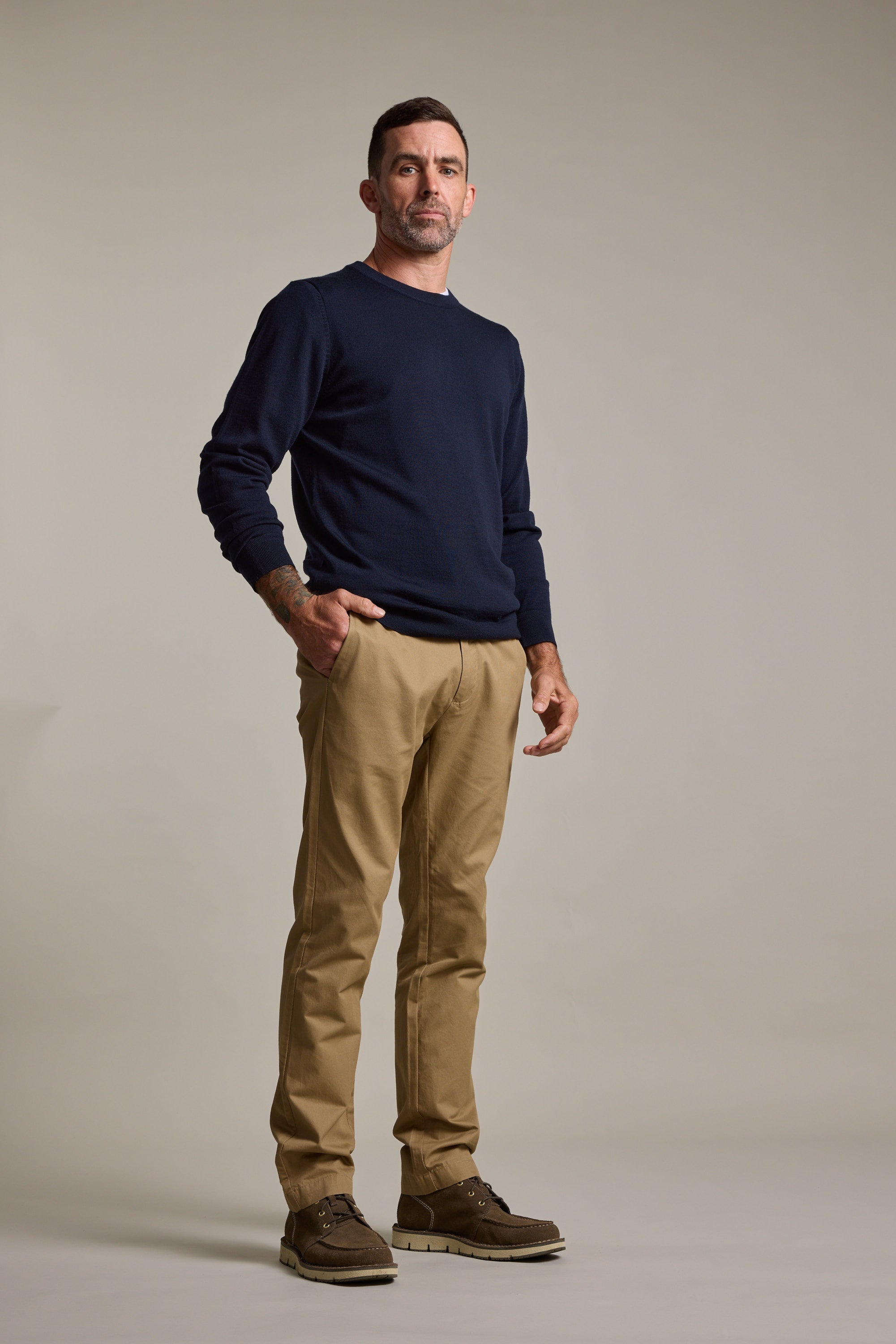 A man with short dark hair and a beard stands in a studio, wearing Barkers Dundas Chino trousers, a navy sweater, and brown shoes. He has a neutral expression, one hand in his pocket, against a plain light background.