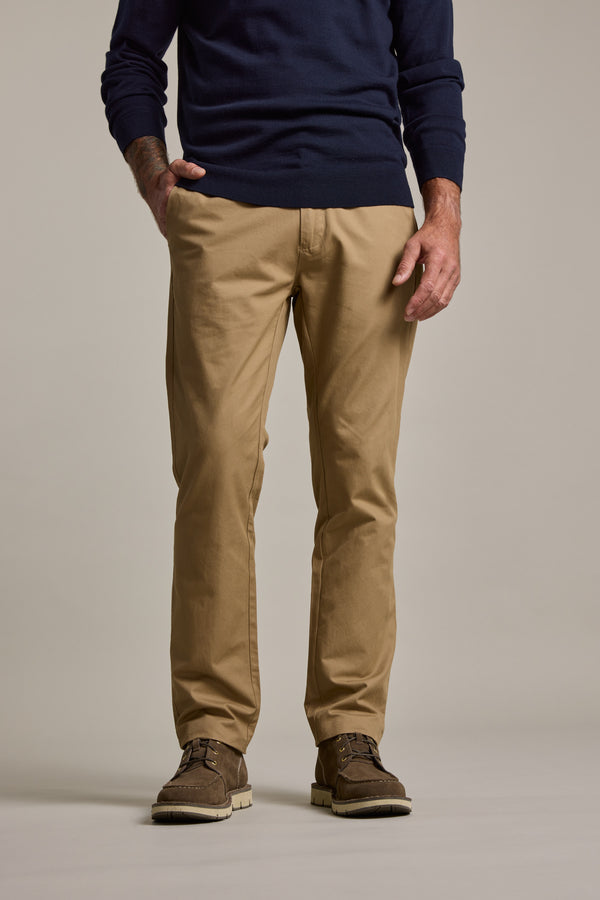 A person stands with one hand in their pocket, wearing Barkers Dundas Chino pants in a relaxed fit, a navy sweater, and brown shoes. The image is cropped from the shoulders down against a plain background.