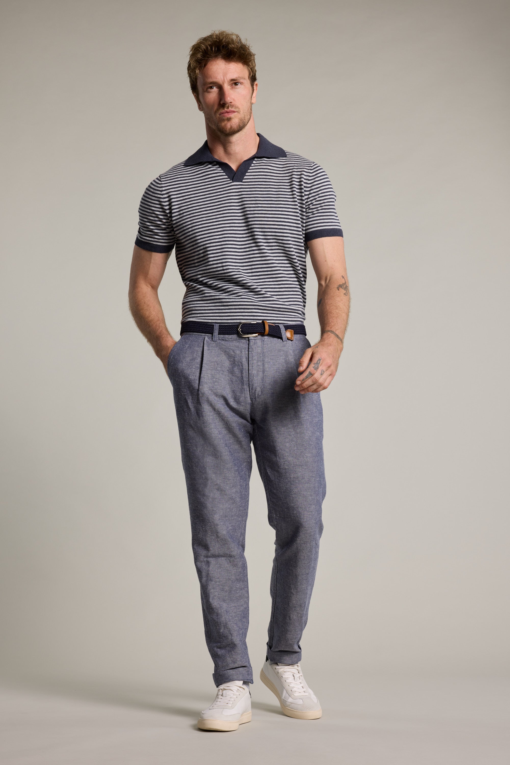 A man with short hair and a trimmed beard poses with his hands in his pockets, wearing the Barkers Parkway Cotton Stripe Polo, blue pleated belted trousers, and white sneakers against a plain background.
