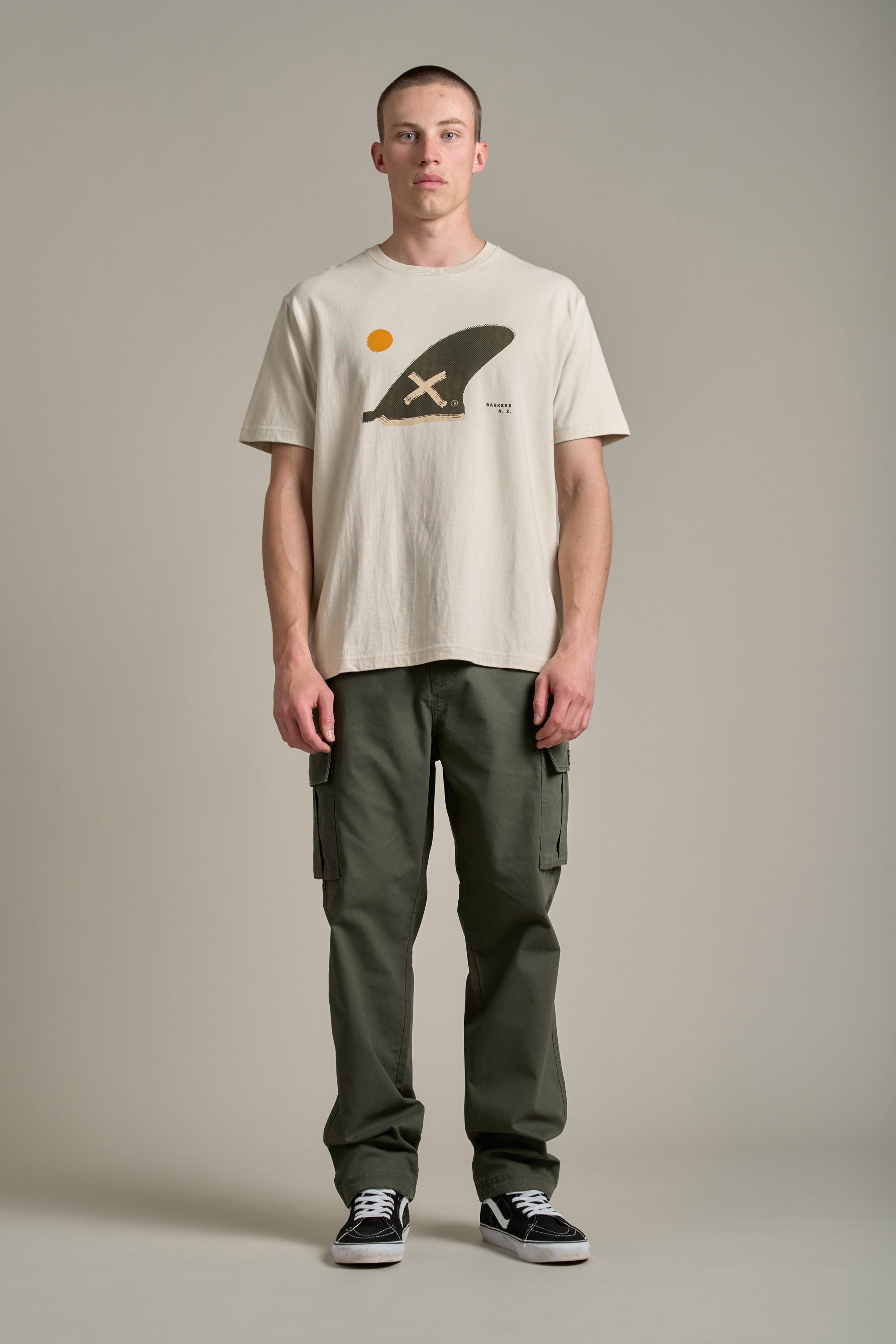 A man stands against a plain background wearing a beige graphic helmet tee, Barkers Randall Cargo Pants in olive green twill cotton for utilitarian style, and black sneakers. He faces forward with a neutral expression and hands at his sides.