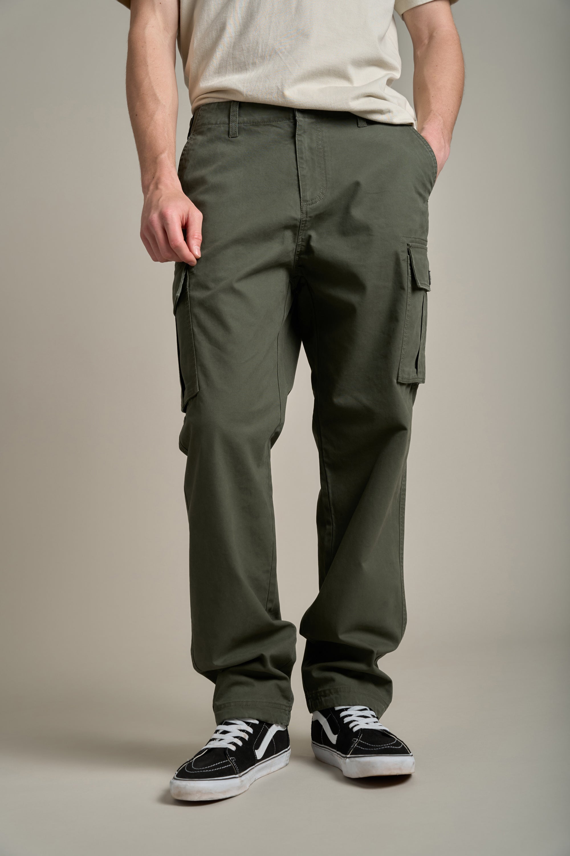 A person stands against a plain background wearing Barkers Randall Cargo Pants in olive green twill cotton, an off-white t-shirt, and black-and-white sneakers. Hands in pockets highlight the casual utilitarian style; face is not visible.