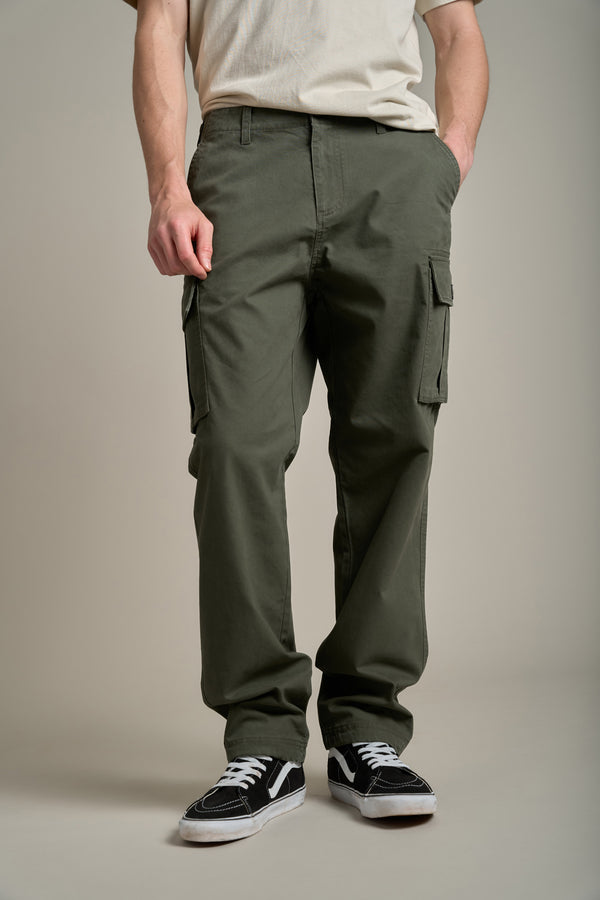 A person stands against a plain background wearing Barkers Randall Cargo Pants in olive green twill cotton, an off-white t-shirt, and black-and-white sneakers. Hands in pockets highlight the casual utilitarian style; face is not visible.