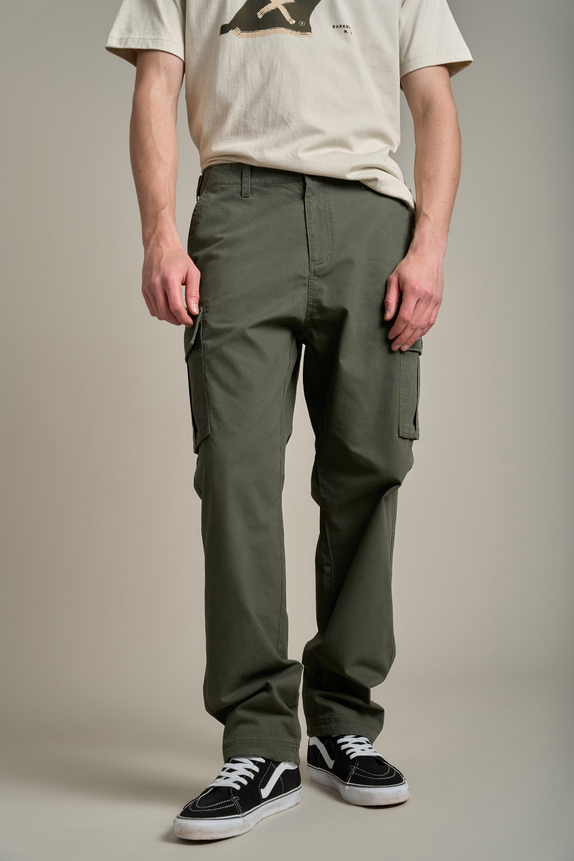 A person stands against a plain background wearing Barkers Randall Cargo Pants in olive green twill cotton, a beige graphic t-shirt, and black low-top sneakers. The utilitarian outfit is modeled with the face not visible.