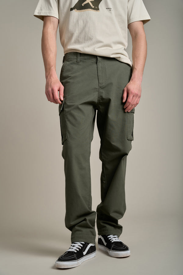 A person stands against a plain background wearing Barkers Randall Cargo Pants in olive green twill cotton, a beige graphic t-shirt, and black low-top sneakers. The utilitarian outfit is modeled with the face not visible.