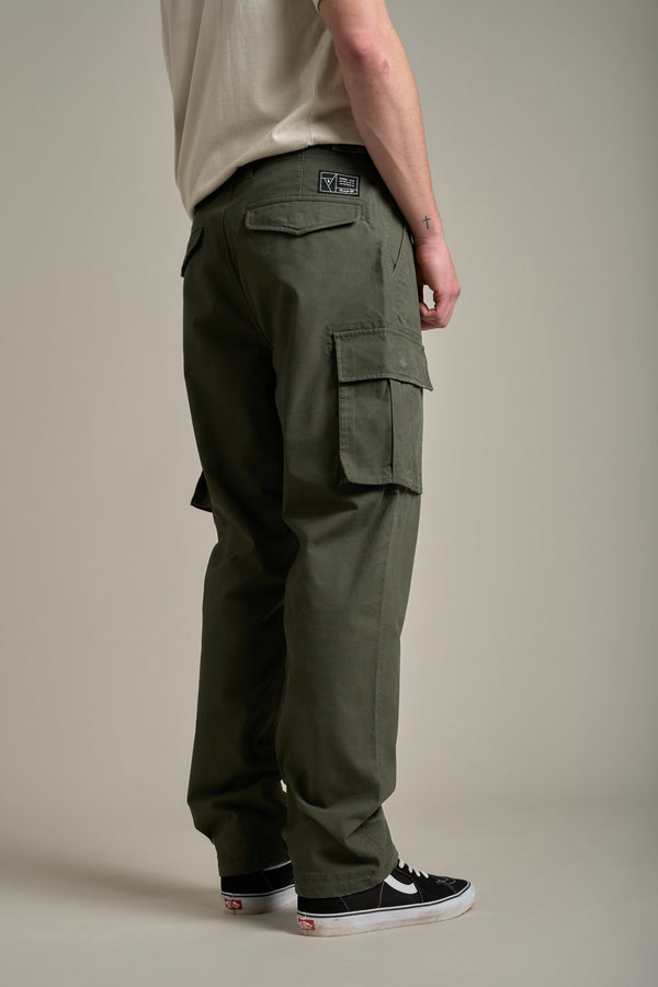 A person stands facing away, wearing Barkers Randall Cargo Pants in olive green twill cotton with side pockets, a beige T-shirt, and black Vans sneakers with white soles, creating a relaxed utilitarian look against a plain background.
