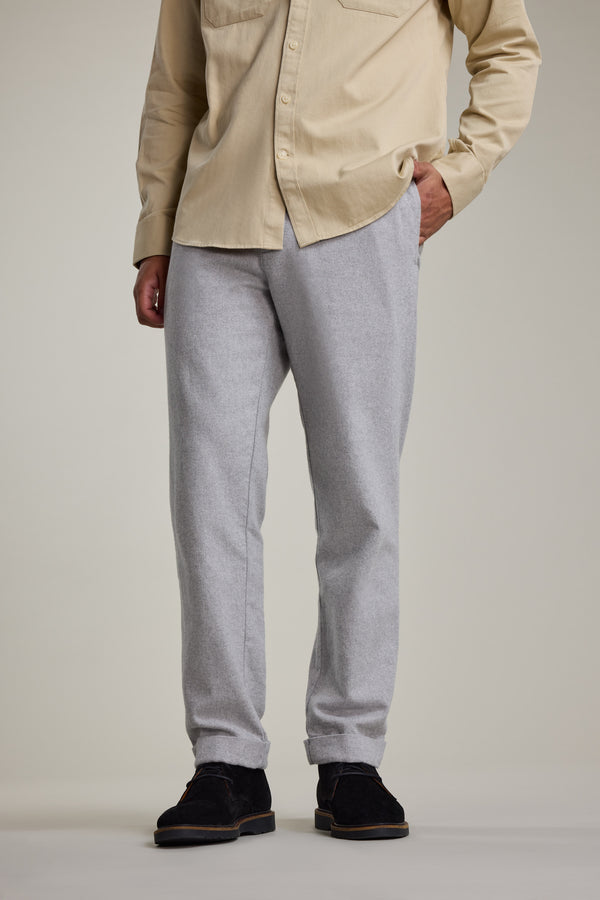 A person stands with one hand in their pocket, wearing Barkers Dunmore Pants in light gray, a beige button-up shirt, and black shoes. Their head and upper torso are out of frame against a plain background.