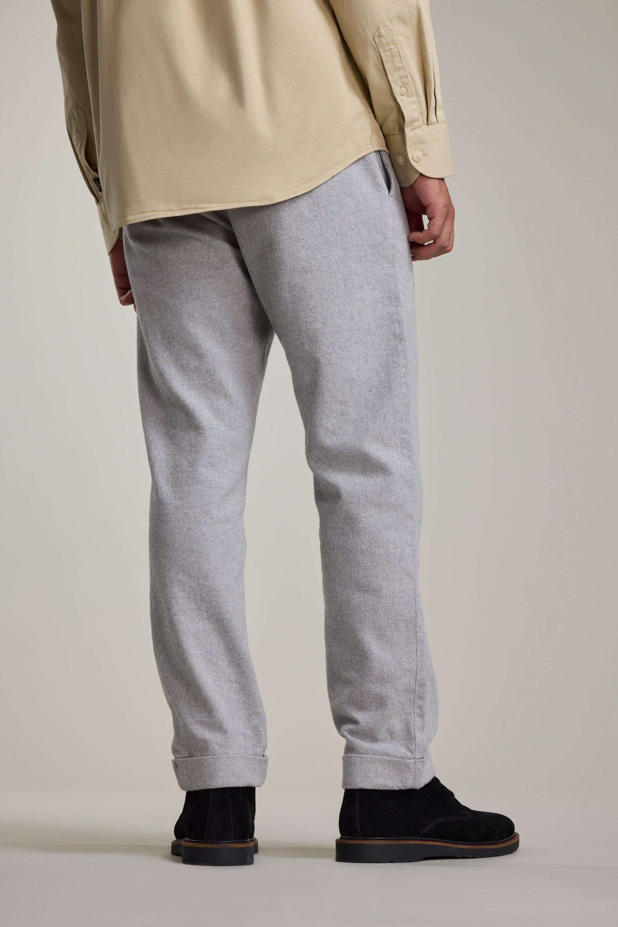 A person stands with their back to the camera, wearing Barkers' Dunmore Pant in relaxed fit, paired with a beige long-sleeve shirt and black shoes. The pants are made from responsibly sourced cotton, shown against a plain neutral background.