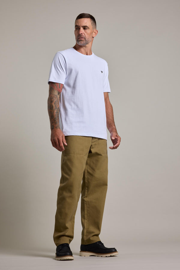 A man with short hair and tattoos on his right arm stands against a plain background, wearing Barkers Midwood Fatique Pant, a white t-shirt, and black shoes. He looks to his left with a neutral expression.