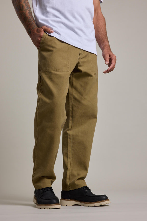 Someone stands against a plain background with one hand in their pocket, wearing Barkers' Midwood Fatique Pant in khaki, a white shirt, and black work shoes with light-colored soles.
