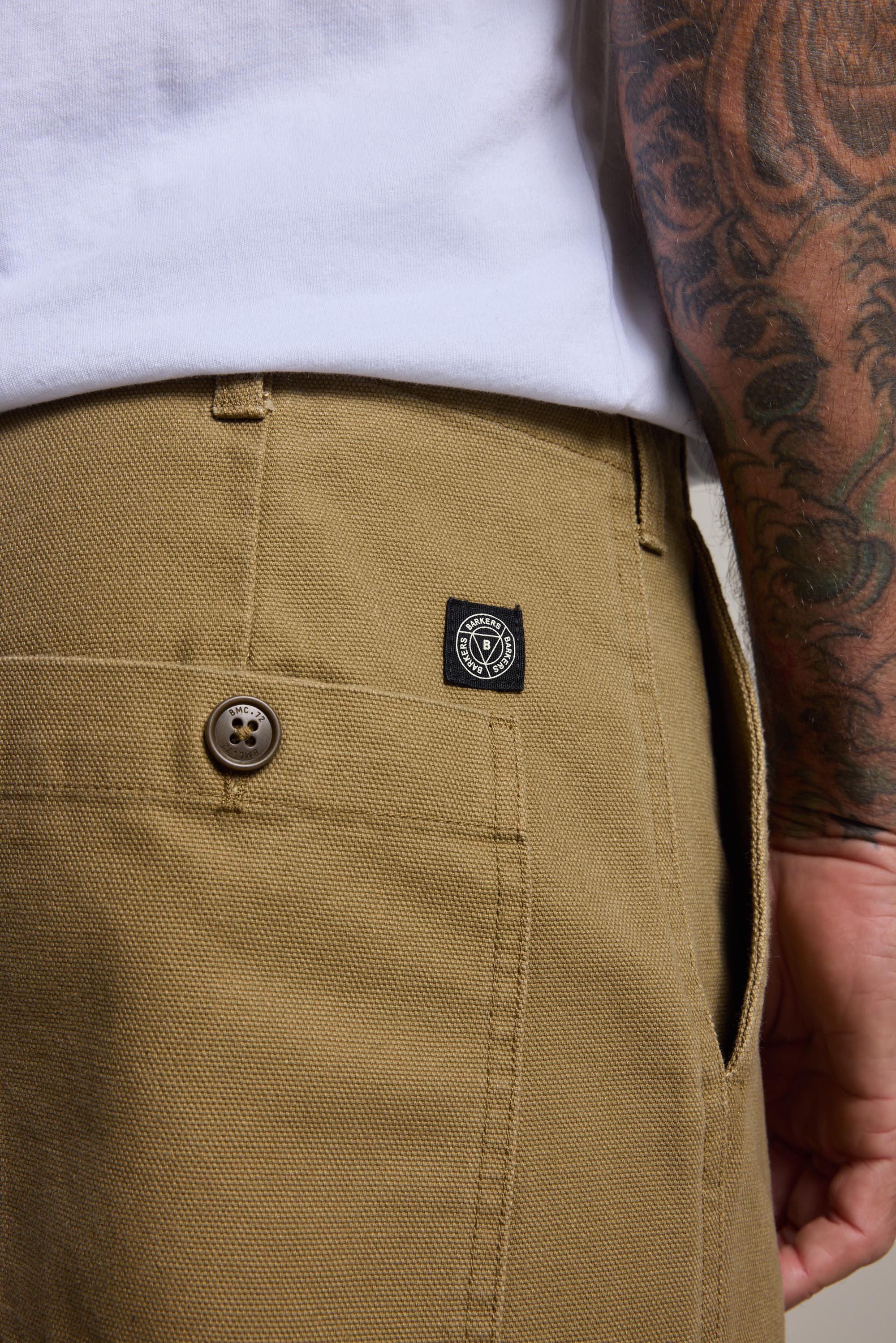 Close-up of a person wearing Barkers Midwood Fatigue Pant in khaki, featuring a relaxed fit, buttoned back pocket, and black logo tag. Styled with a white shirt and showcasing a tattooed arm resting by their side.