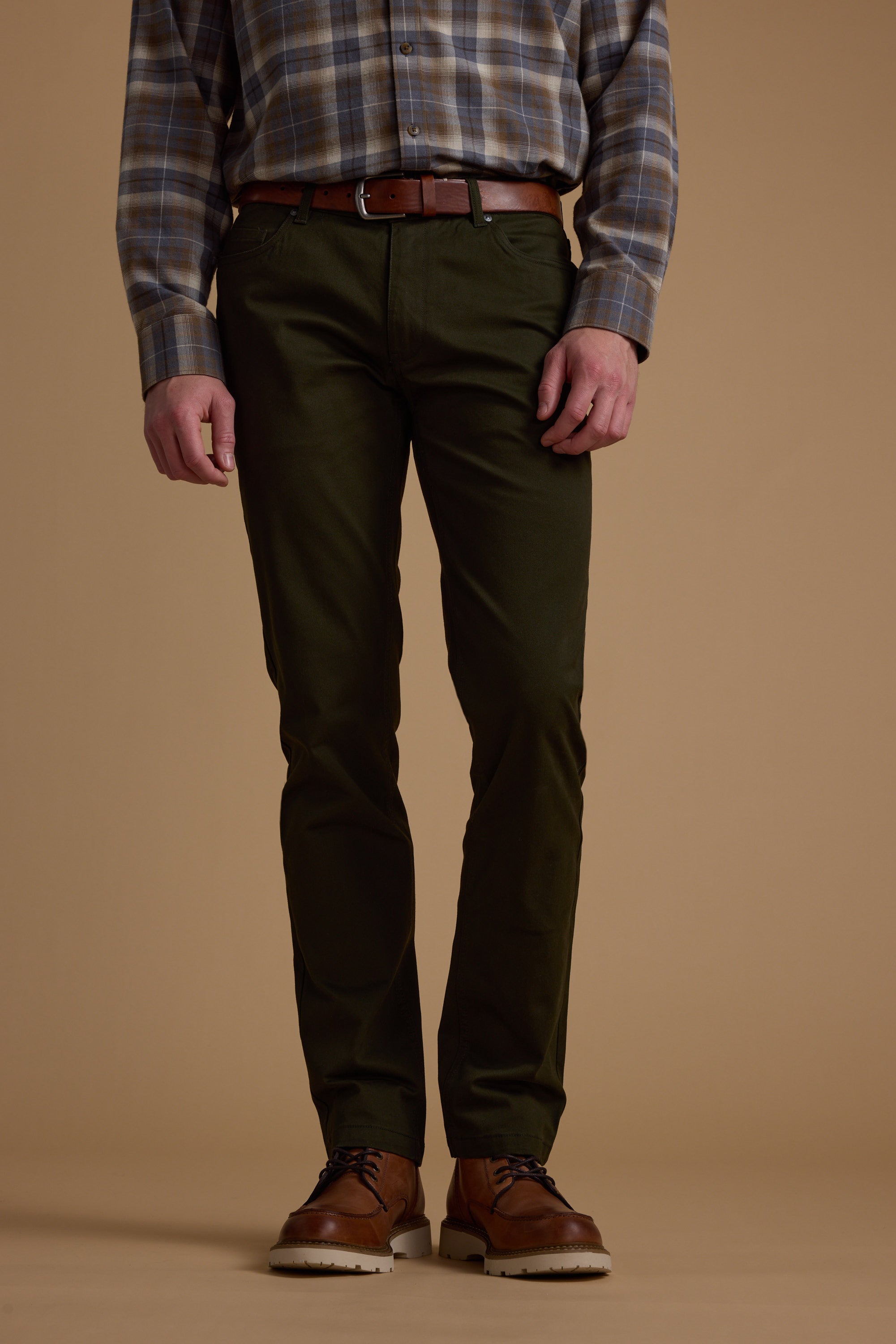 A person stands against a beige background wearing Barkers Beckett 5-Pocket Pant in dark green, paired with a plaid shirt, brown belt, and brown leather shoes. The photo is cropped from the shoulders down.