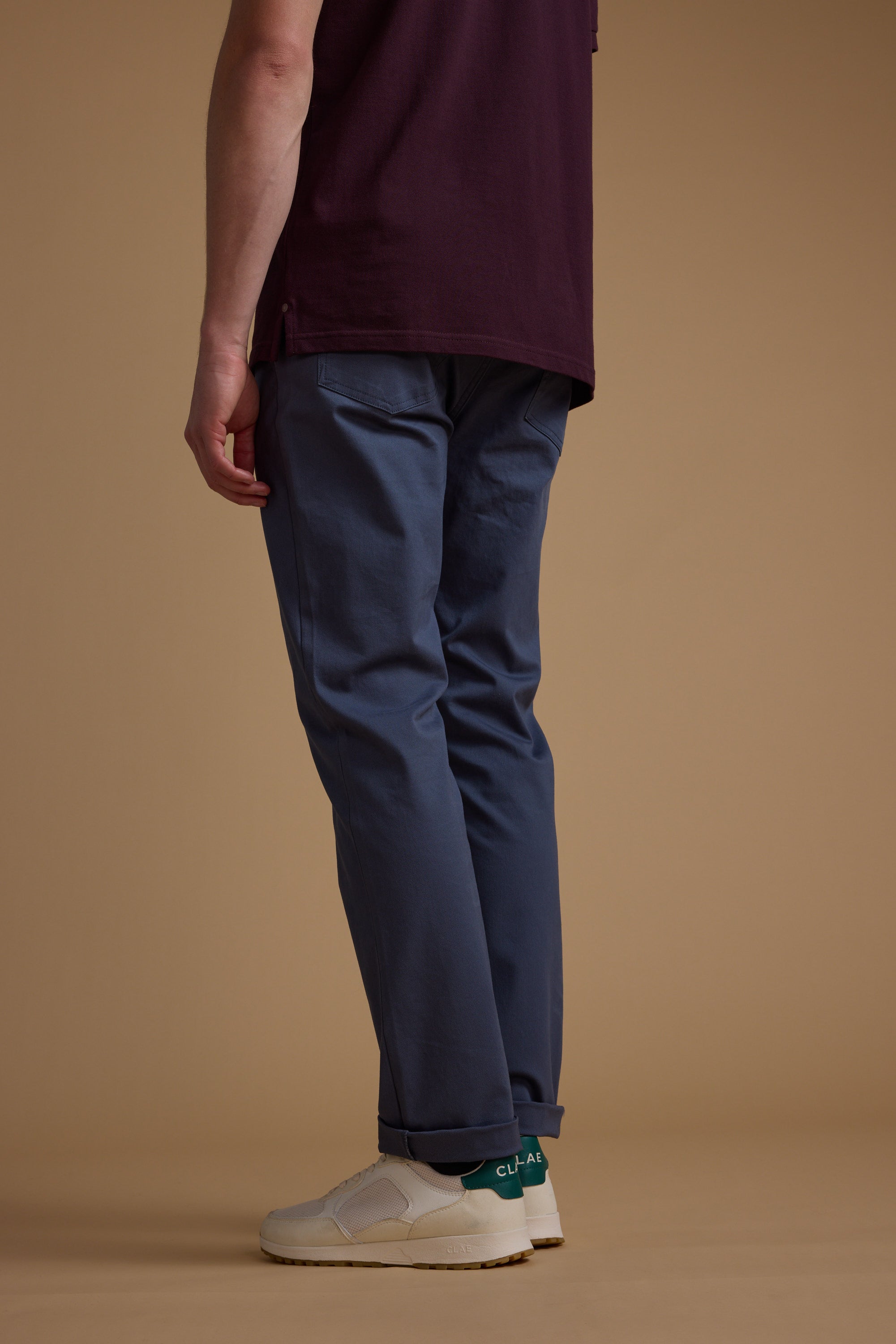 Against a beige background, a person wears Barkers Beckett 5-Pocket Pant, styled with a dark purple shirt and white sneakers. The image is cropped at the shoulders to highlight the lower half of the body.