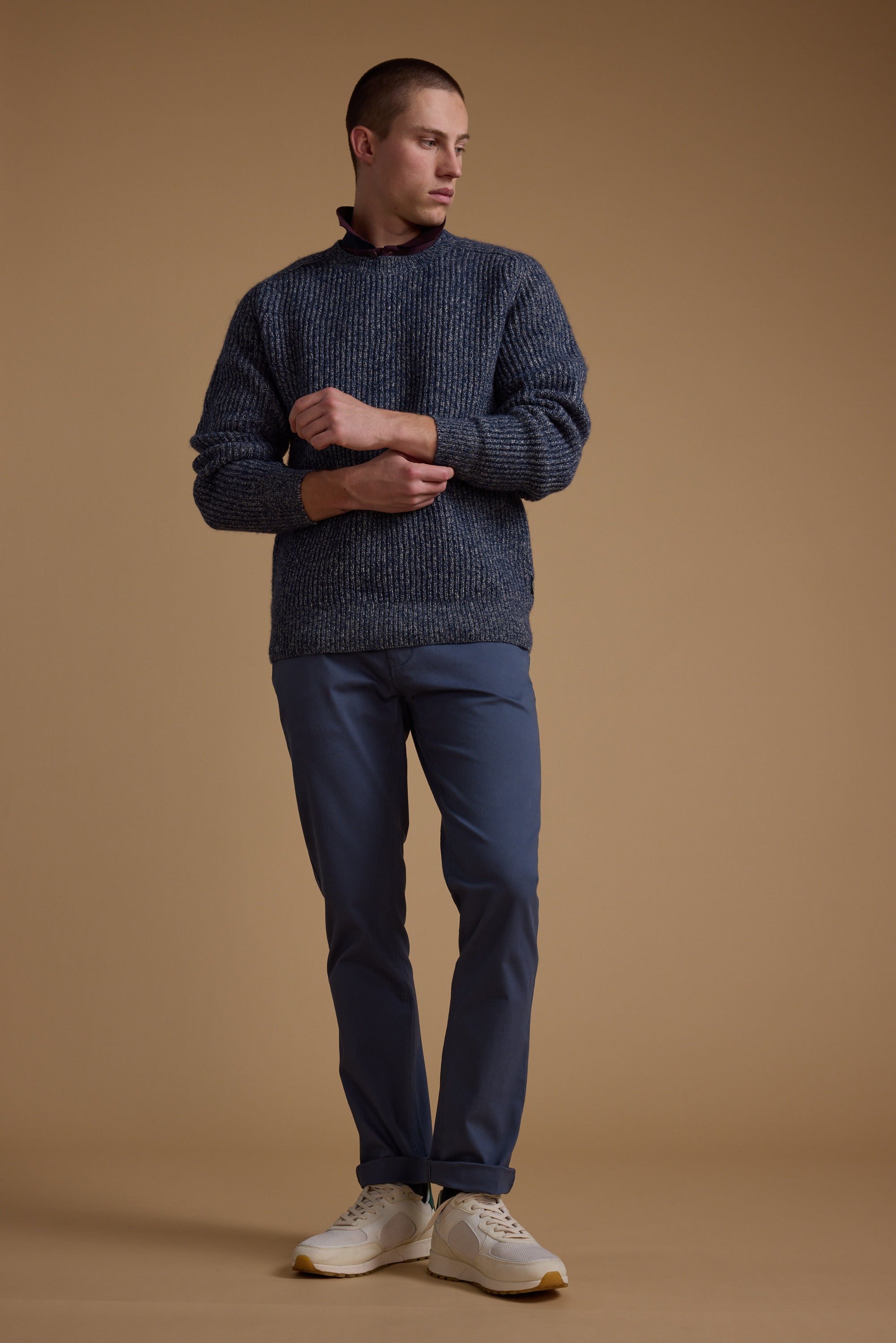 A person stands against a plain brown background, wearing Barkers Beckett 5-Pocket Pant with a chunky blue-gray sweater and white sneakers. They have short hair, look to the side, and adjust one sleeve.