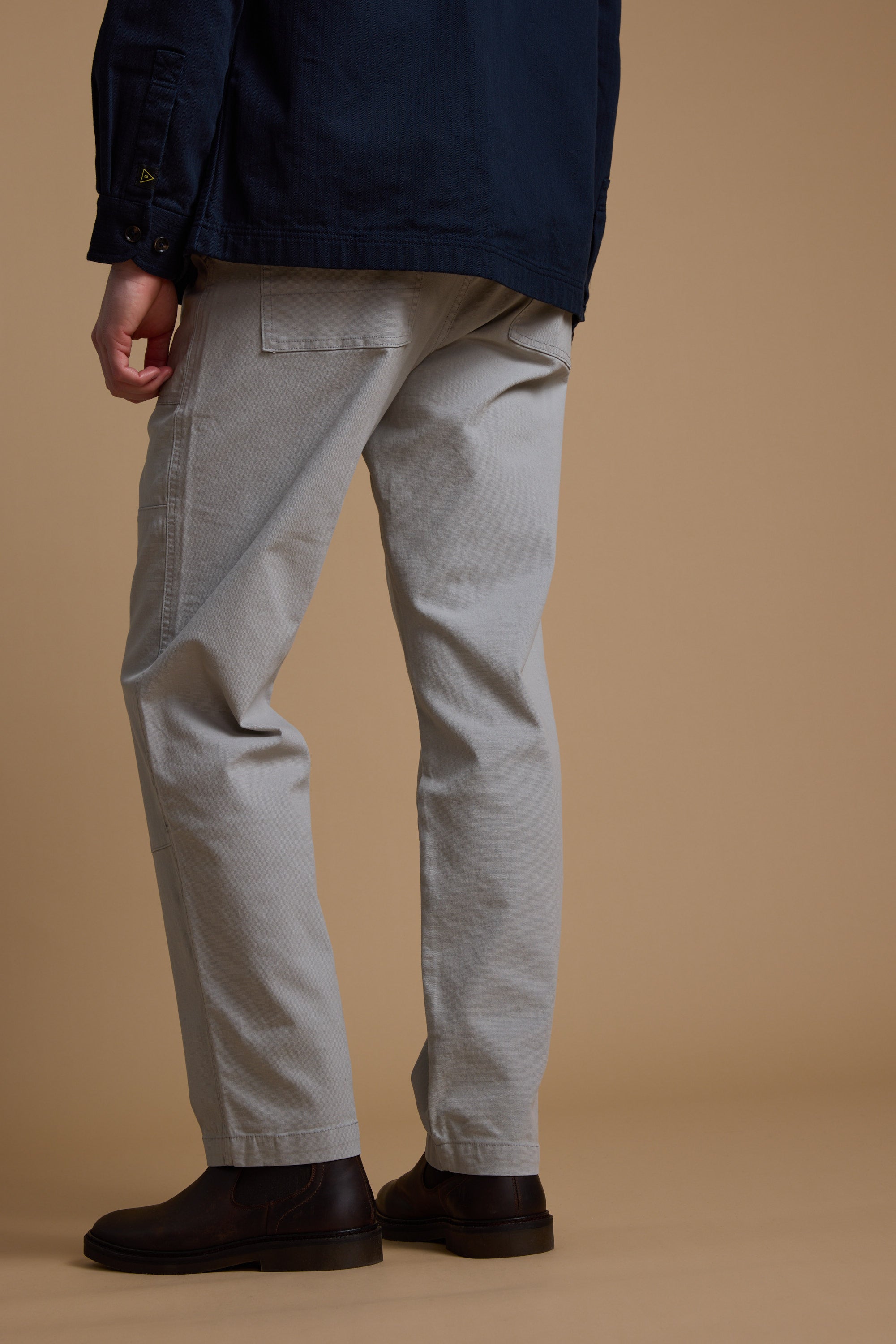 A person stands with their back to the camera, showing only the lower half of their body, wearing Barkers Tidal GD Utility Pant in light grey against a plain beige backdrop.
