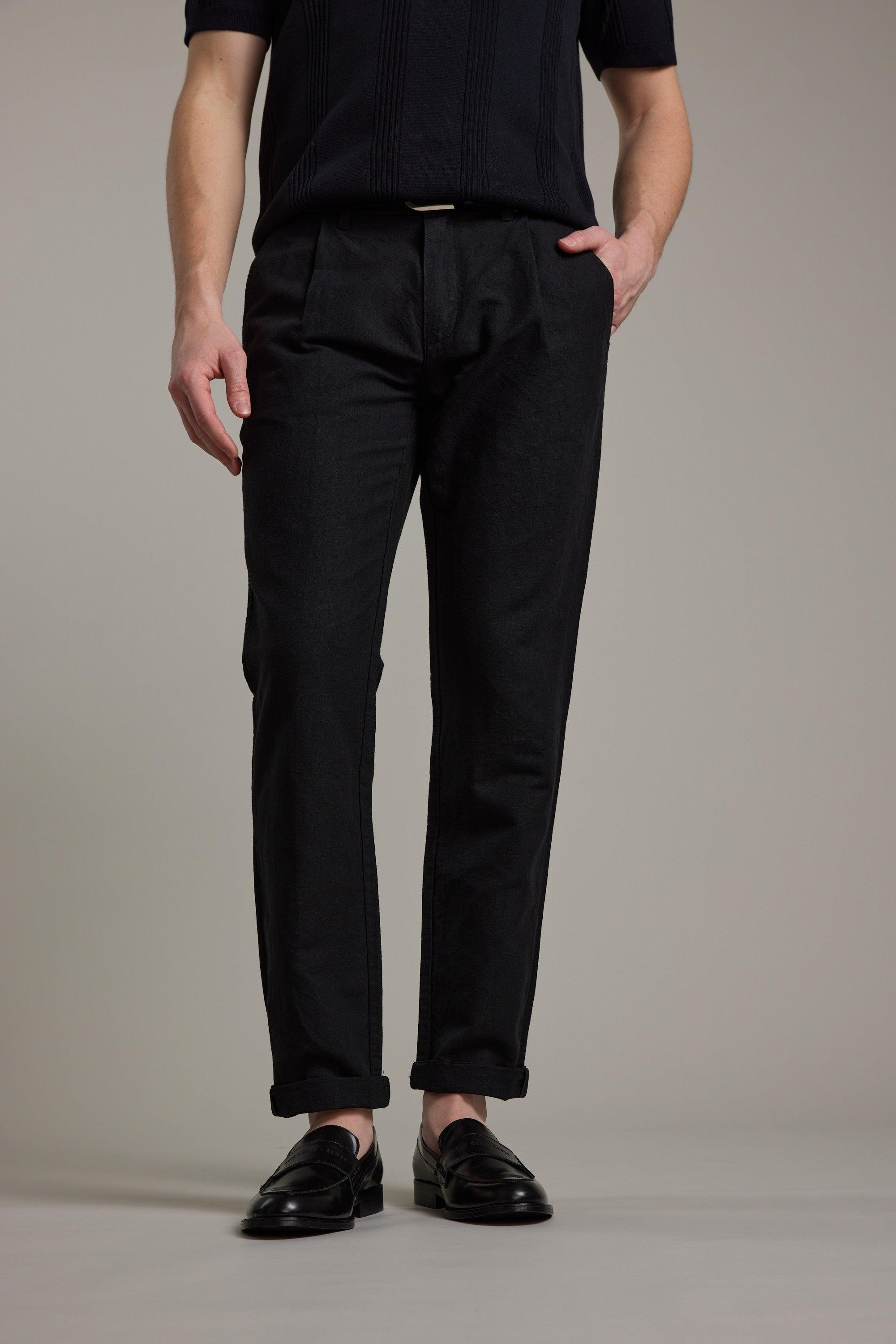 A person stands against a plain background, wearing Barkers' Newman Linen Pant in black with cuffed hems, styled smart-casually with a matching black shirt and loafers. One hand is in their pocket, the other rests relaxed by their side.