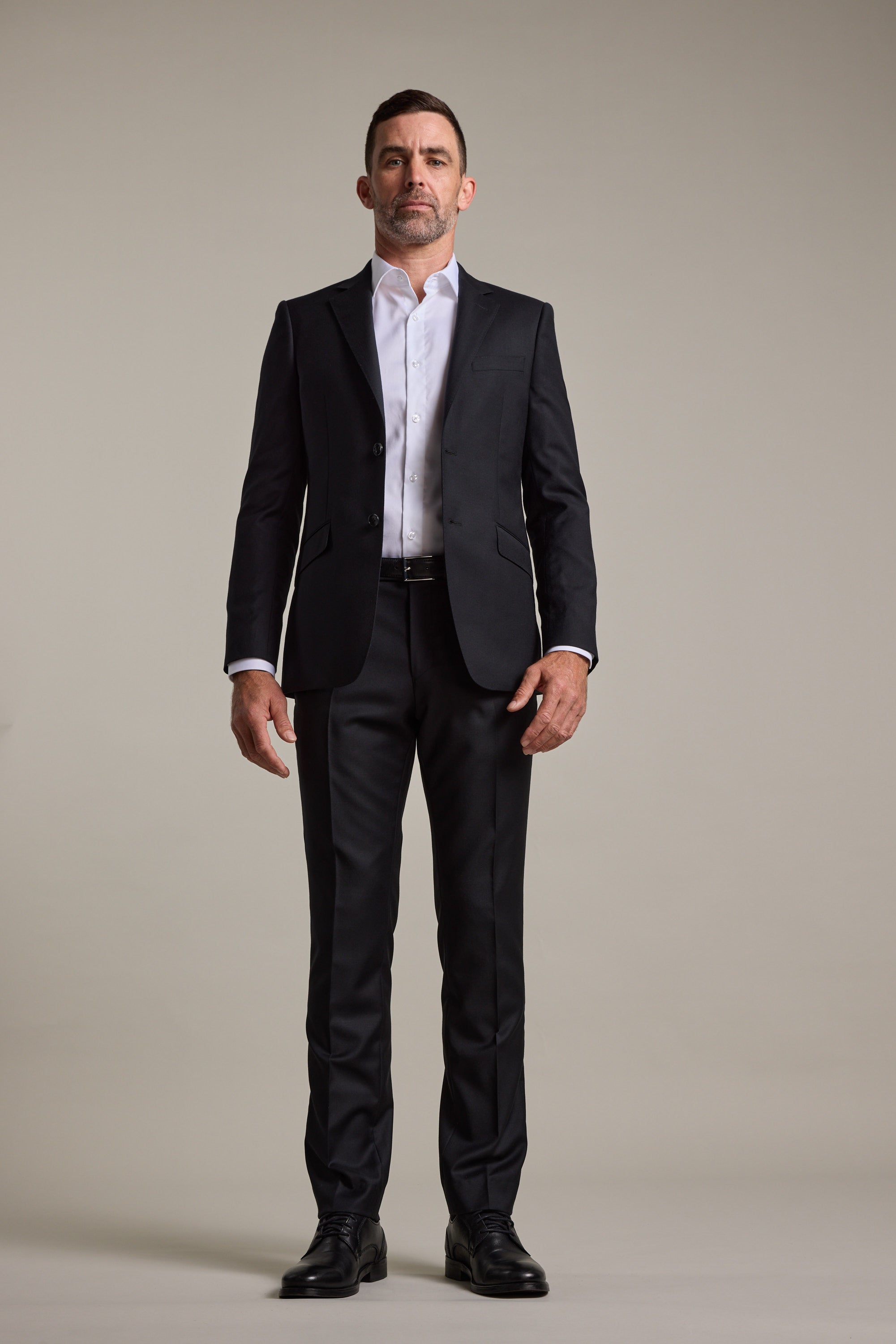 A man wears the Barkers Mayfair ZQrx Suit Jacket in sleek black merino wool, paired with a white shirt and black dress shoes, standing against a beige background—an ideal wedding suit look.