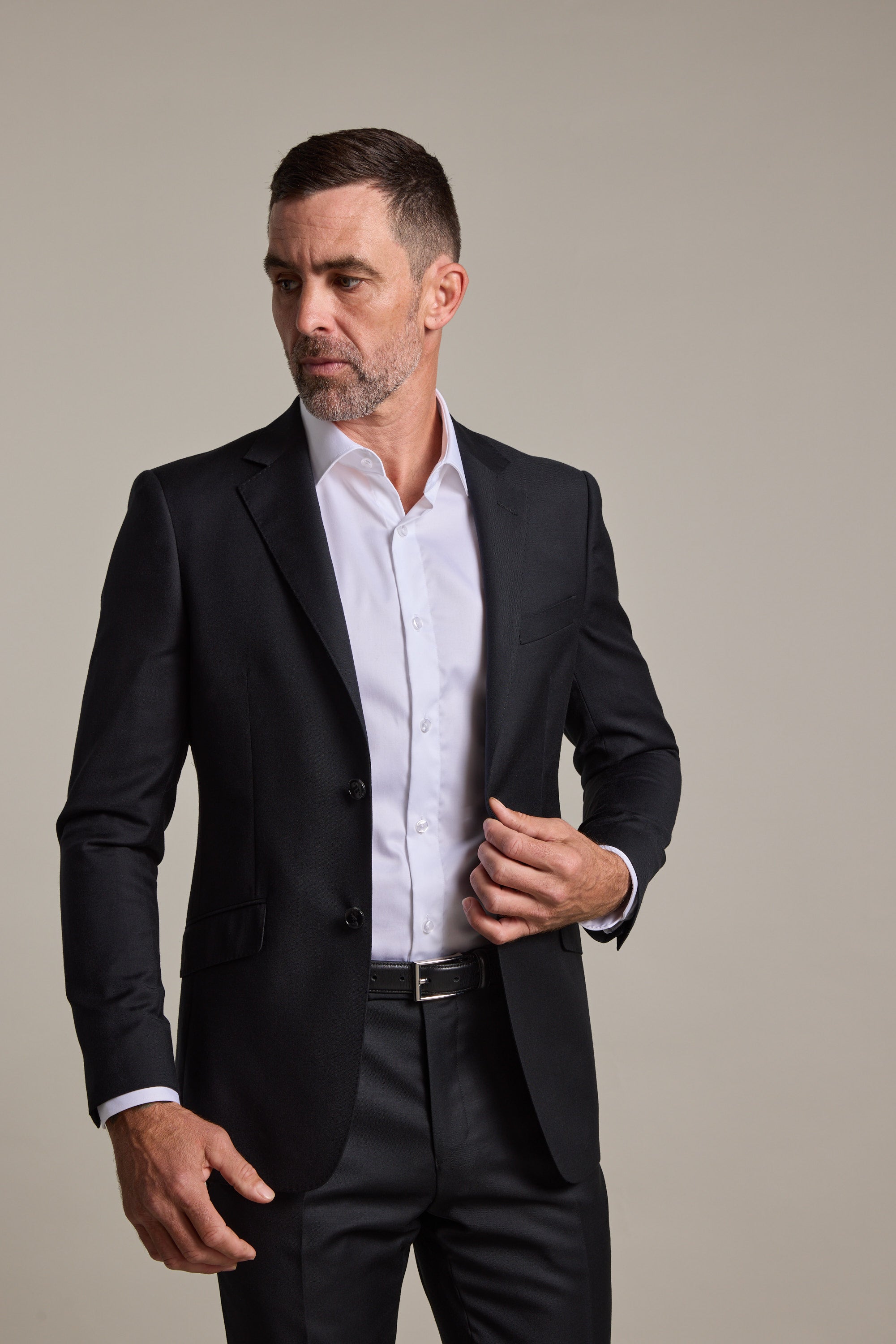 A man with short brown hair and a beard wears the Barkers Mayfair ZQrx Suit Jacket over a white dress shirt. He looks slightly left, adjusting his jacket with his right hand against a plain light background.