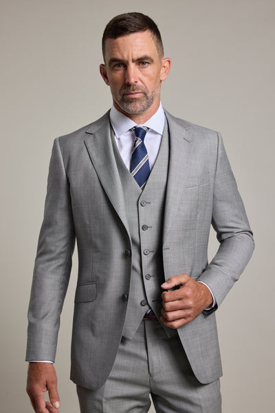 A man with short dark hair and a trimmed beard wears the Barkers Henry Waistcoat as part of a light grey wool three-piece suit, styled with a white shirt and striped navy tie, against a plain light background.