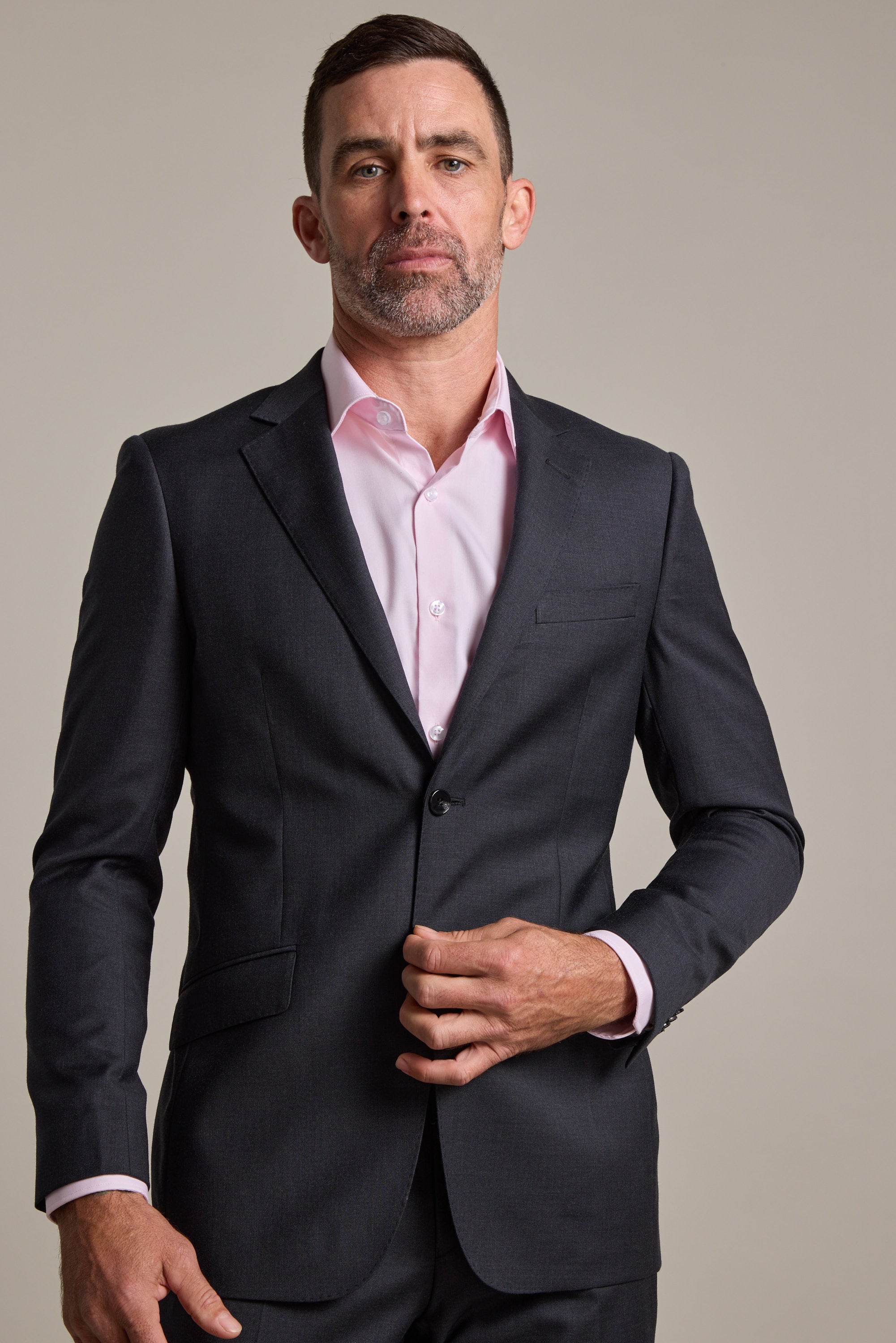 A man with short dark hair and a beard wears a Barkers Mayfair ZQ Suit Jacket and a light pink dress shirt, standing against a plain light background while buttoning his jacket.