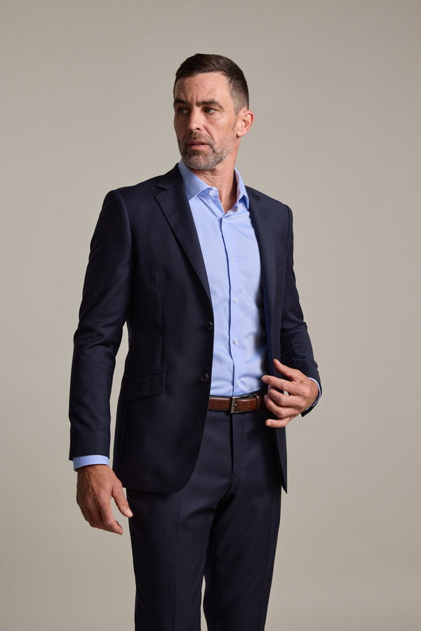 A man with short dark hair and a beard wears a Barkers Mayfair ZQ Suit Jacket in navy with a light blue dress shirt, standing against a plain background. He looks to the side with one hand near his jacket.