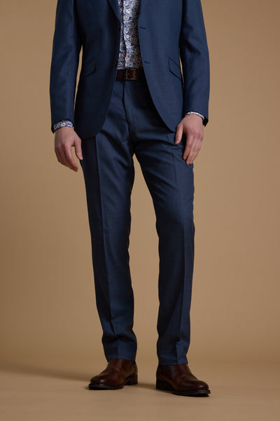 A man, shown from the neck down, models Barkers’ Morden Texture Suit Trouser—men’s blue dress pants—in a tailored fit with a patterned shirt, brown belt, and brown shoes against a plain tan background.
