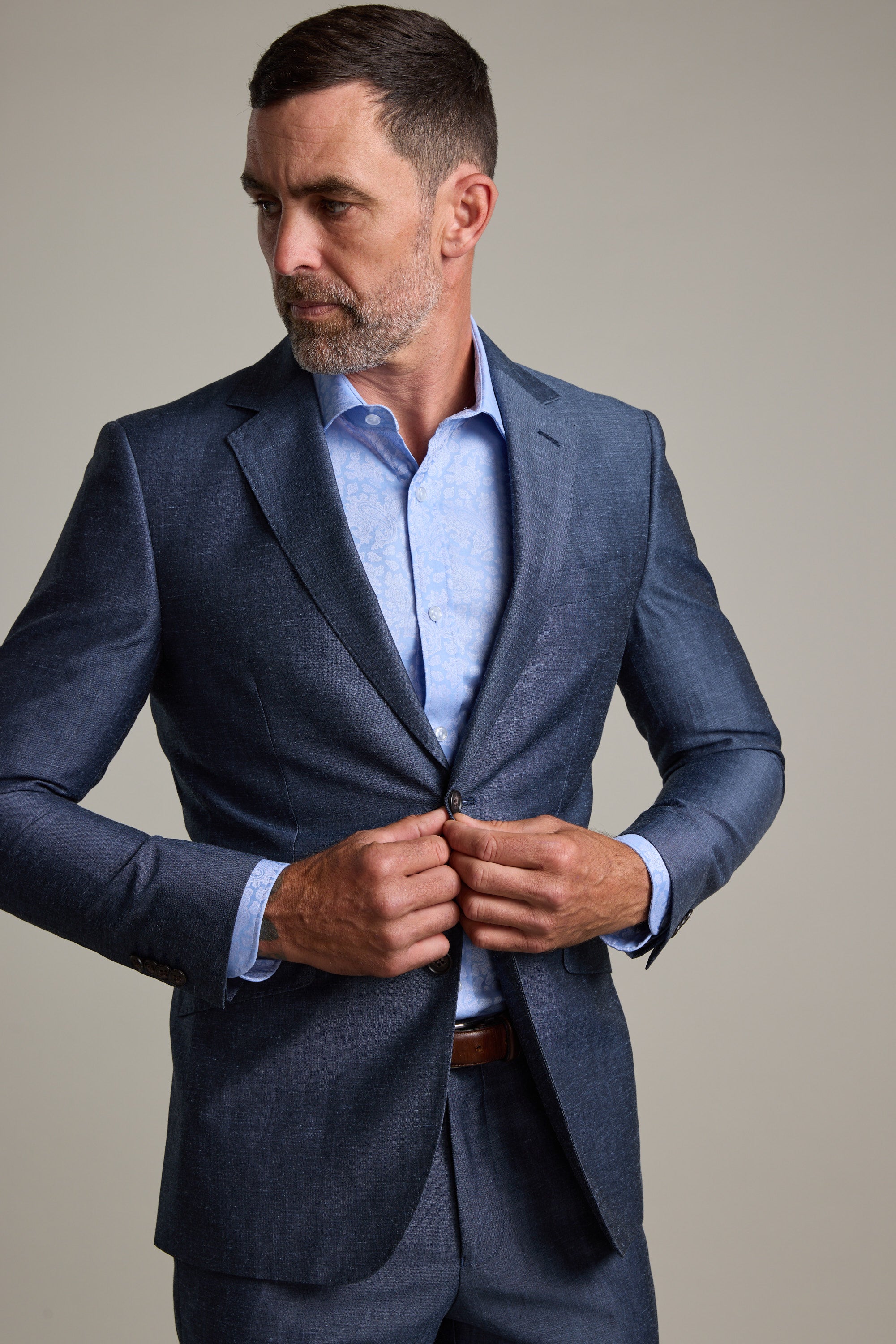 A man with short hair and a beard buttons the Barkers Charles Texture Suit Jacket over a light blue dress shirt, highlighting modern tailoring as he looks to his left against a plain background.