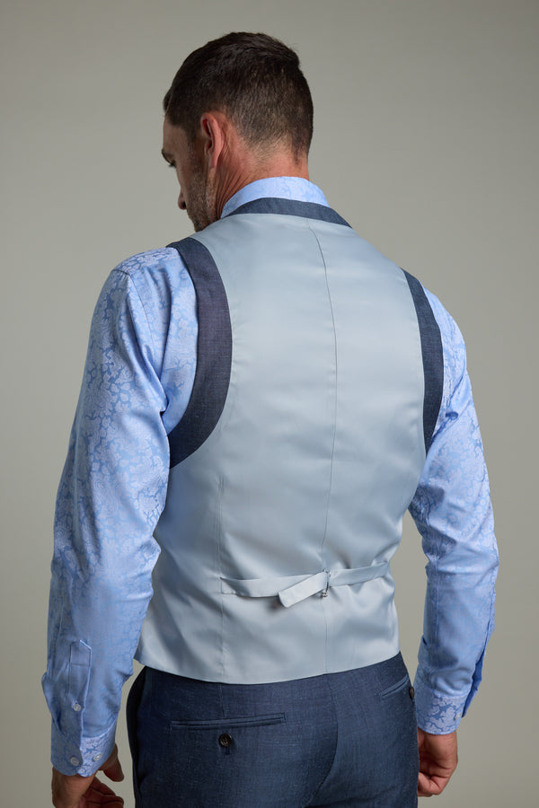 A man is seen from the back in a Barkers Charles Texture Waistcoat with dark trim and adjustable back buckle, layered over a patterned light blue dress shirt and blue trousers against a plain gray background.